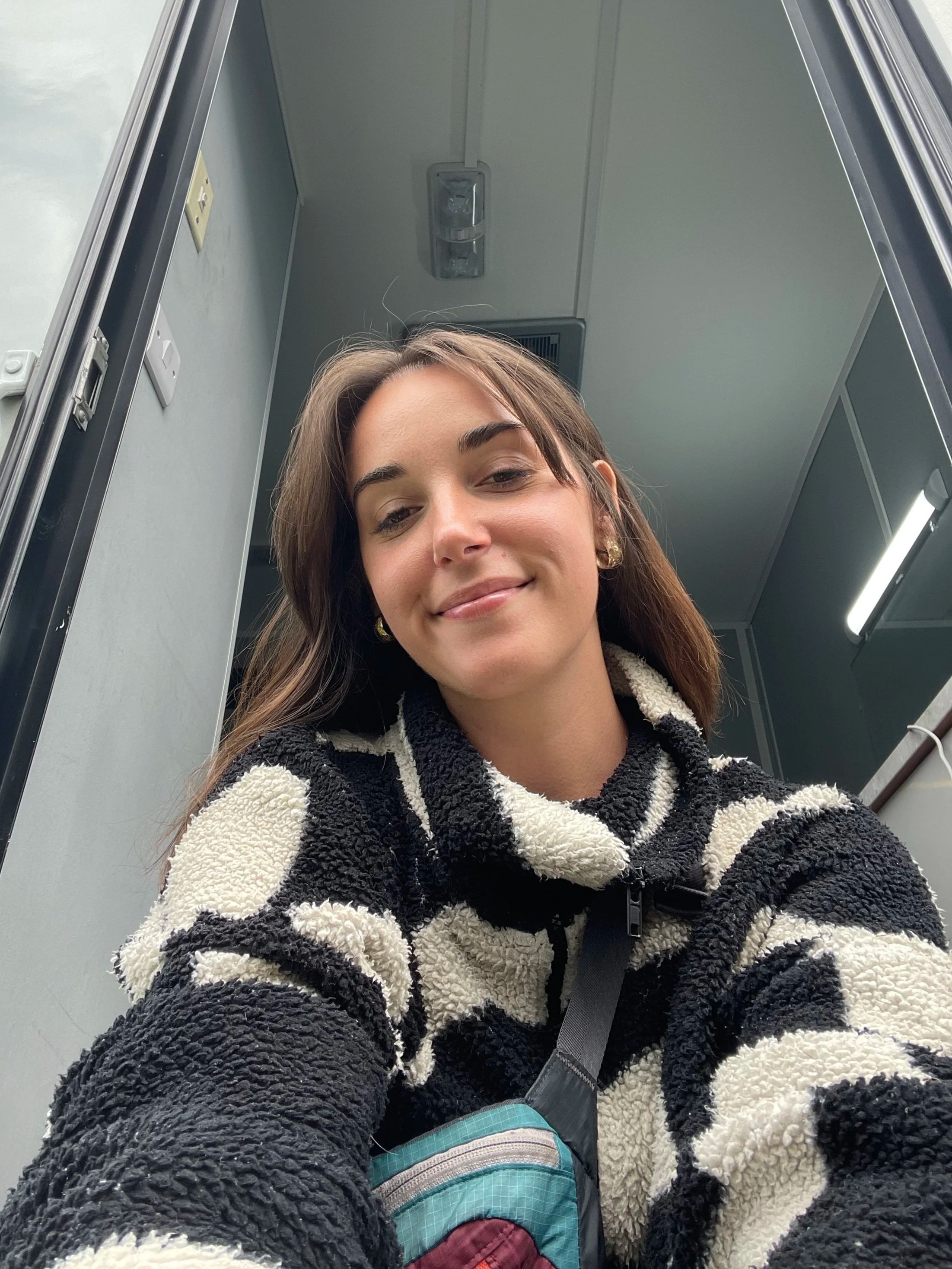 A woman with brown hair and a smile taking a selfie inside a small room or closet, wearing a black and white fuzzy patterned jacket and a seatbelt.