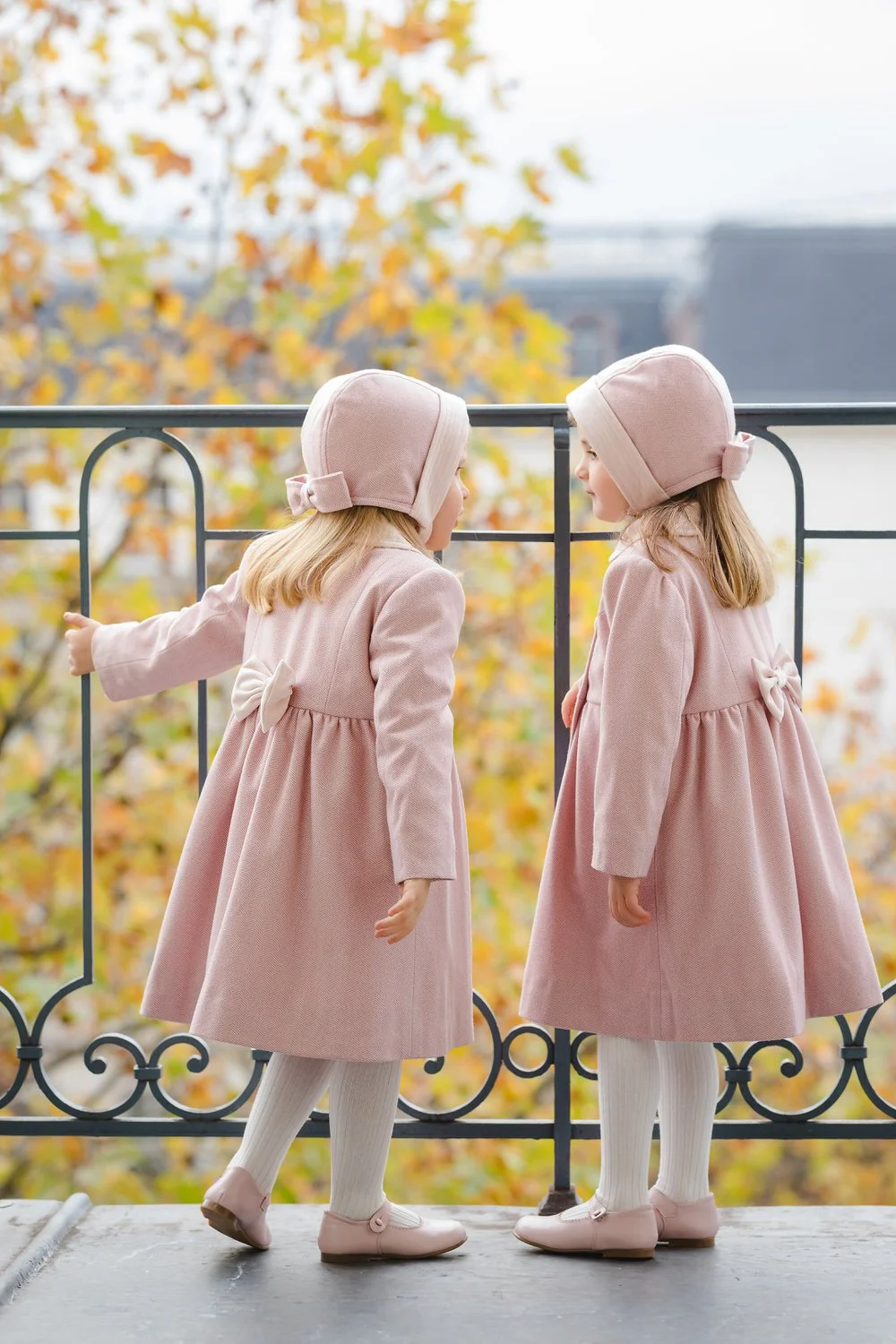 Two young girls dressed in pink coats and matching pink hats, standing on a balcony with a black railing, facing each other and whispering, with autumn-colored leaves in the background.