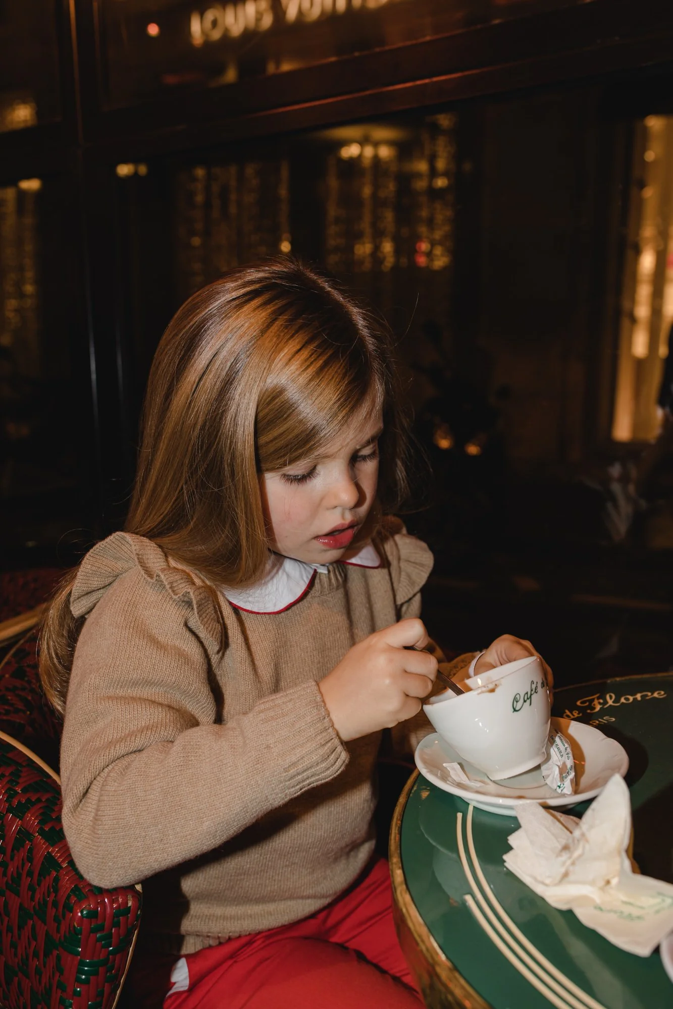 A young girl with light brown hair and a beige sweater sitting at a table in a cafe, stirring a beverage in a white cup.