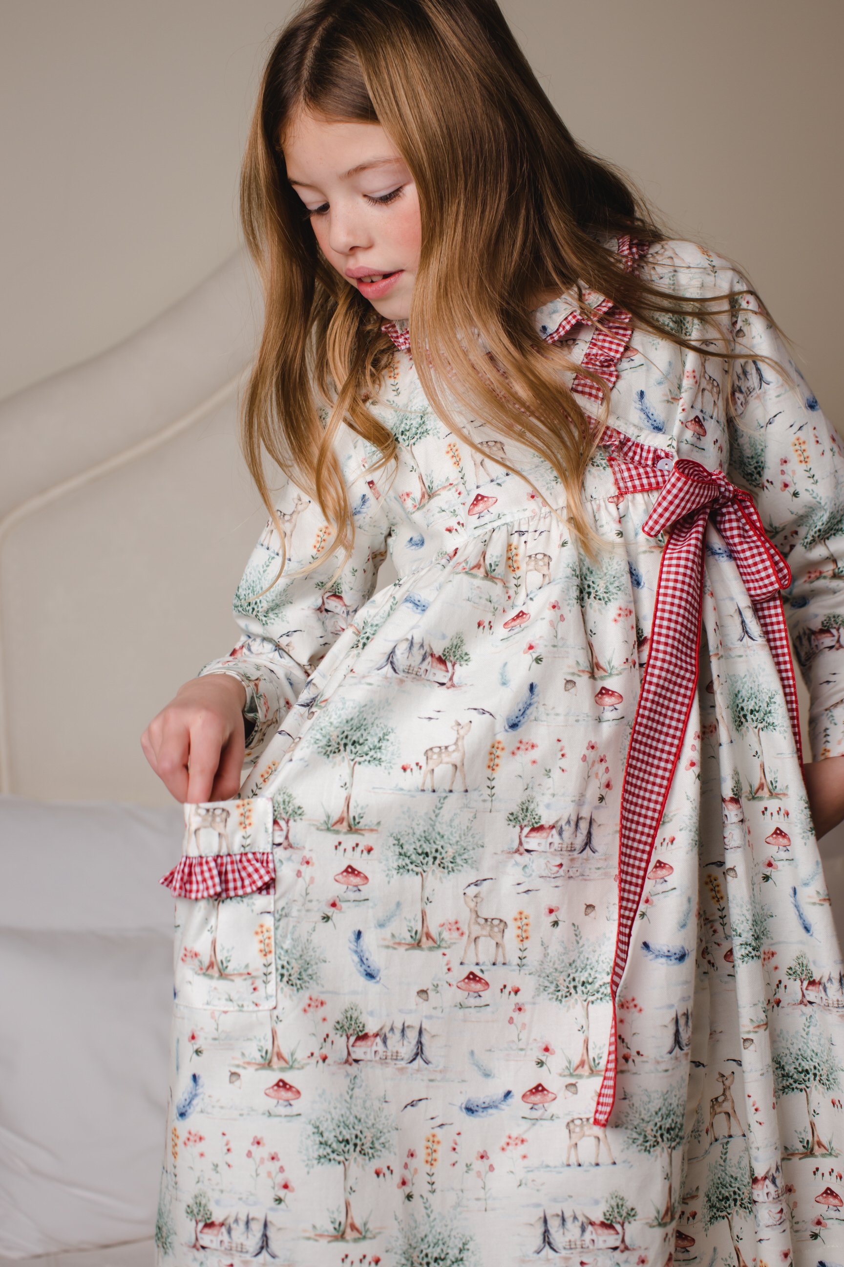 A young girl wearing a long, patterned nightgown with a red checkered ribbon tied at the waist, standing in a bedroom.