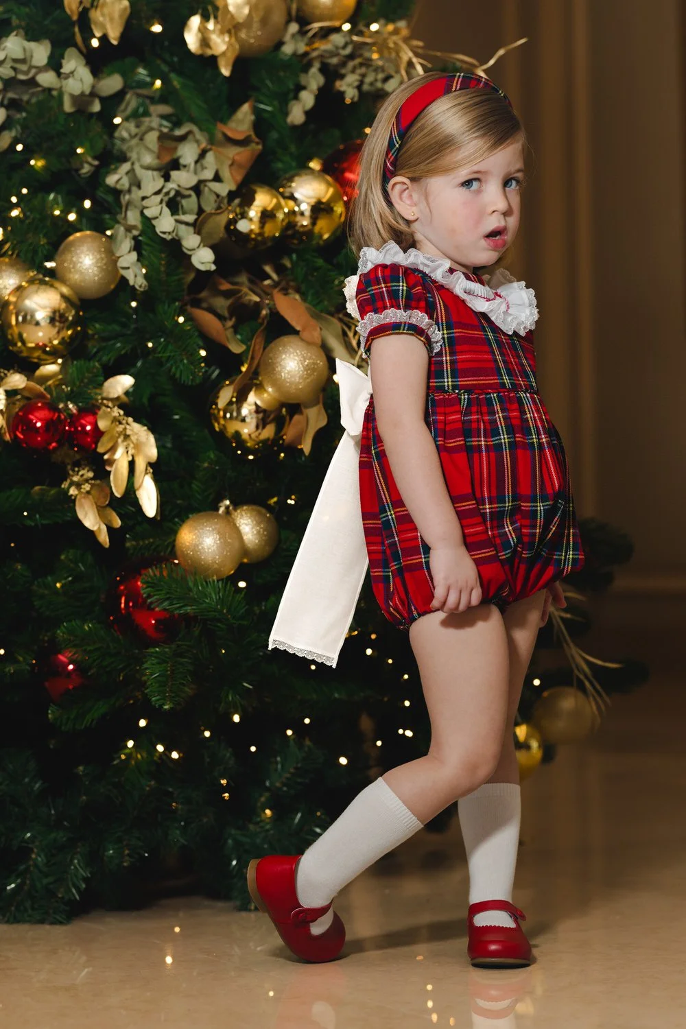 A young girl dressed in a red tartan holiday outfit with a large white bow at the back, standing in front of a decorated Christmas tree with gold and red ornaments and white lights.