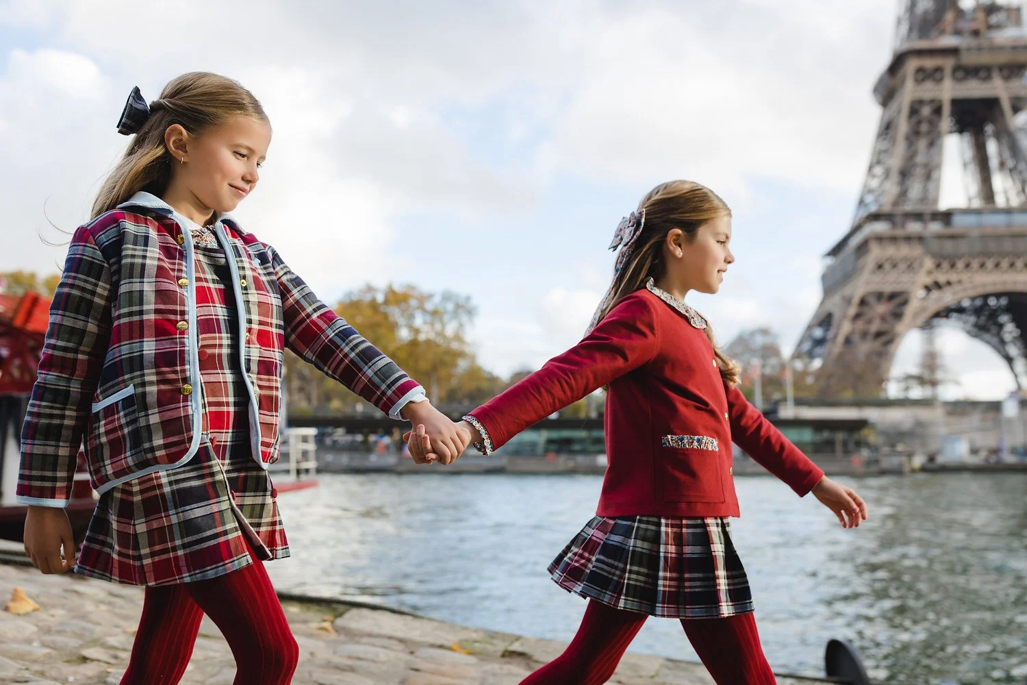 Two girls in red and plaid outfits holding hands and walking along the river in Paris with the Eiffel Tower in the background.