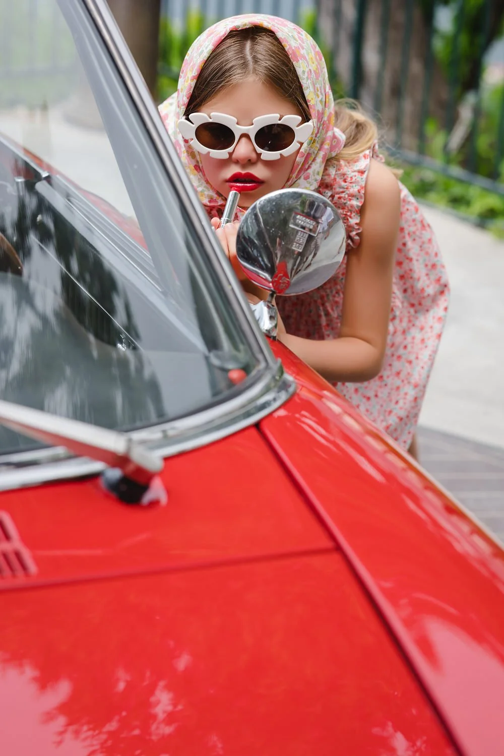 A young woman wearing a floral headscarf and white flower-shaped sunglasses applying lipstick in the side mirror of a red vintage car in Positano.