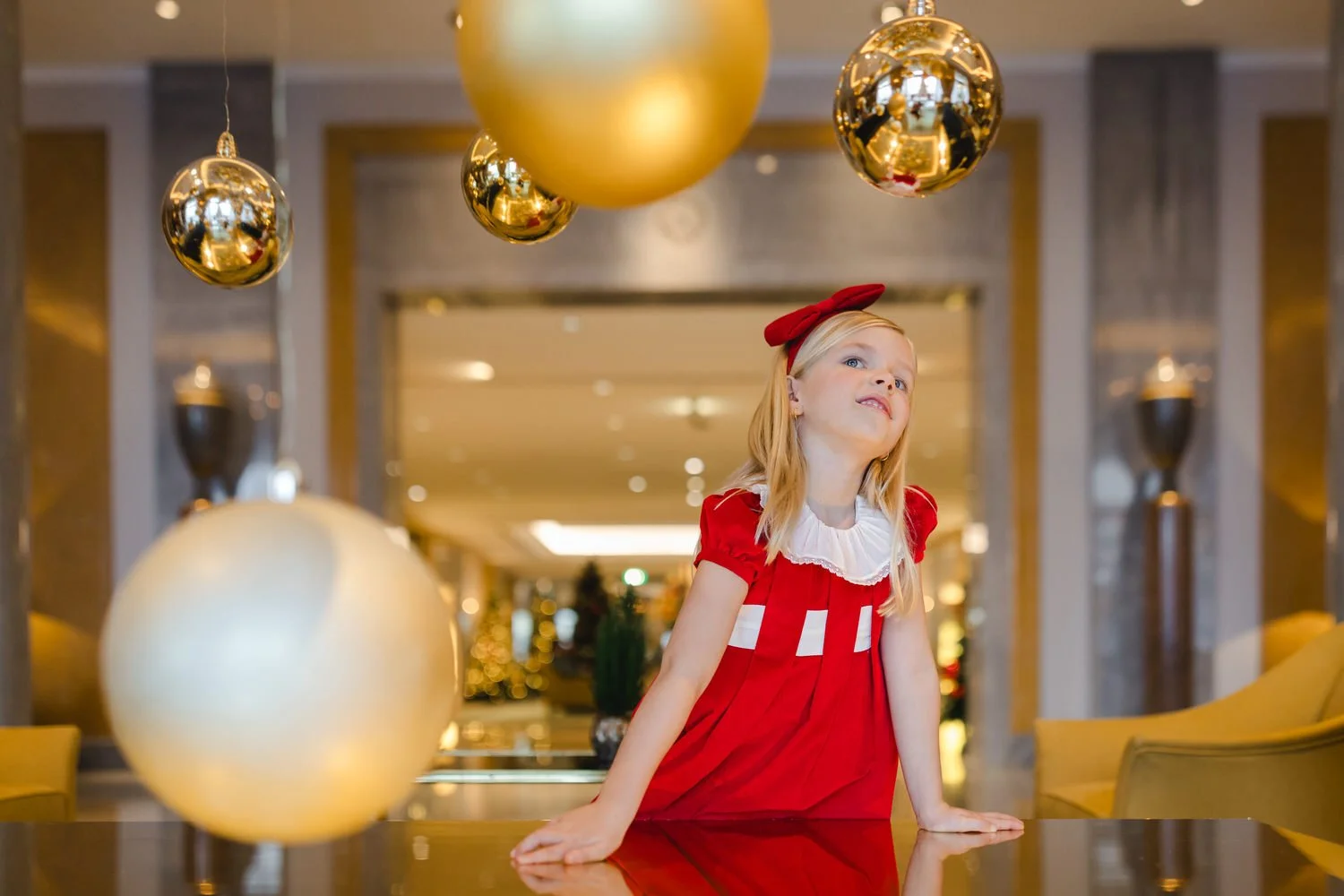 A young girl in a red festive dress with a white collar and a red headband, standing at a table in a decorated, elegant indoor space with gold and black Christmas ornaments hanging above her.