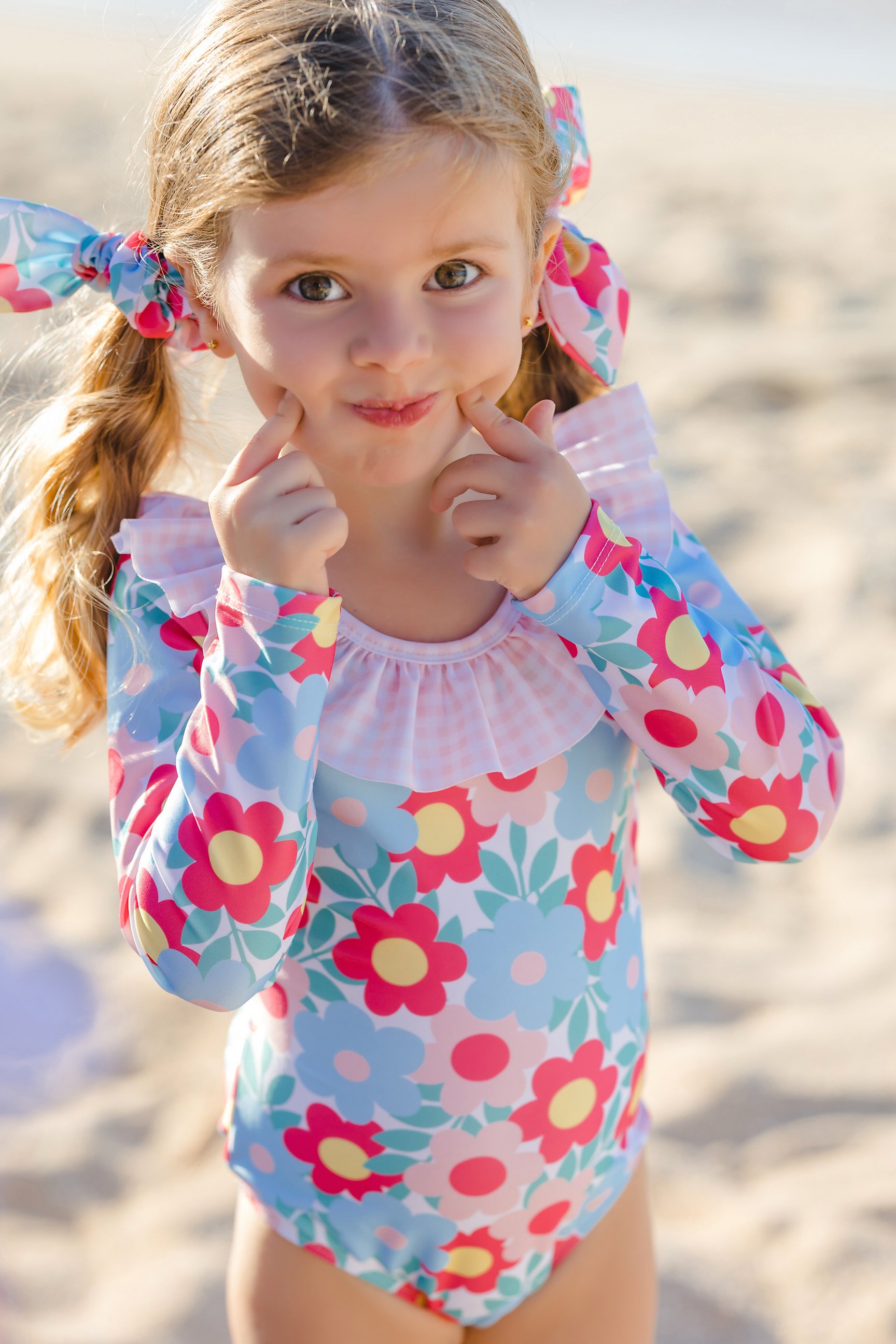 A young girl at the beach with curly hair in pigtails, wearing a colorful floral swimsuit, smiling and touching her cheeks with her fingers.