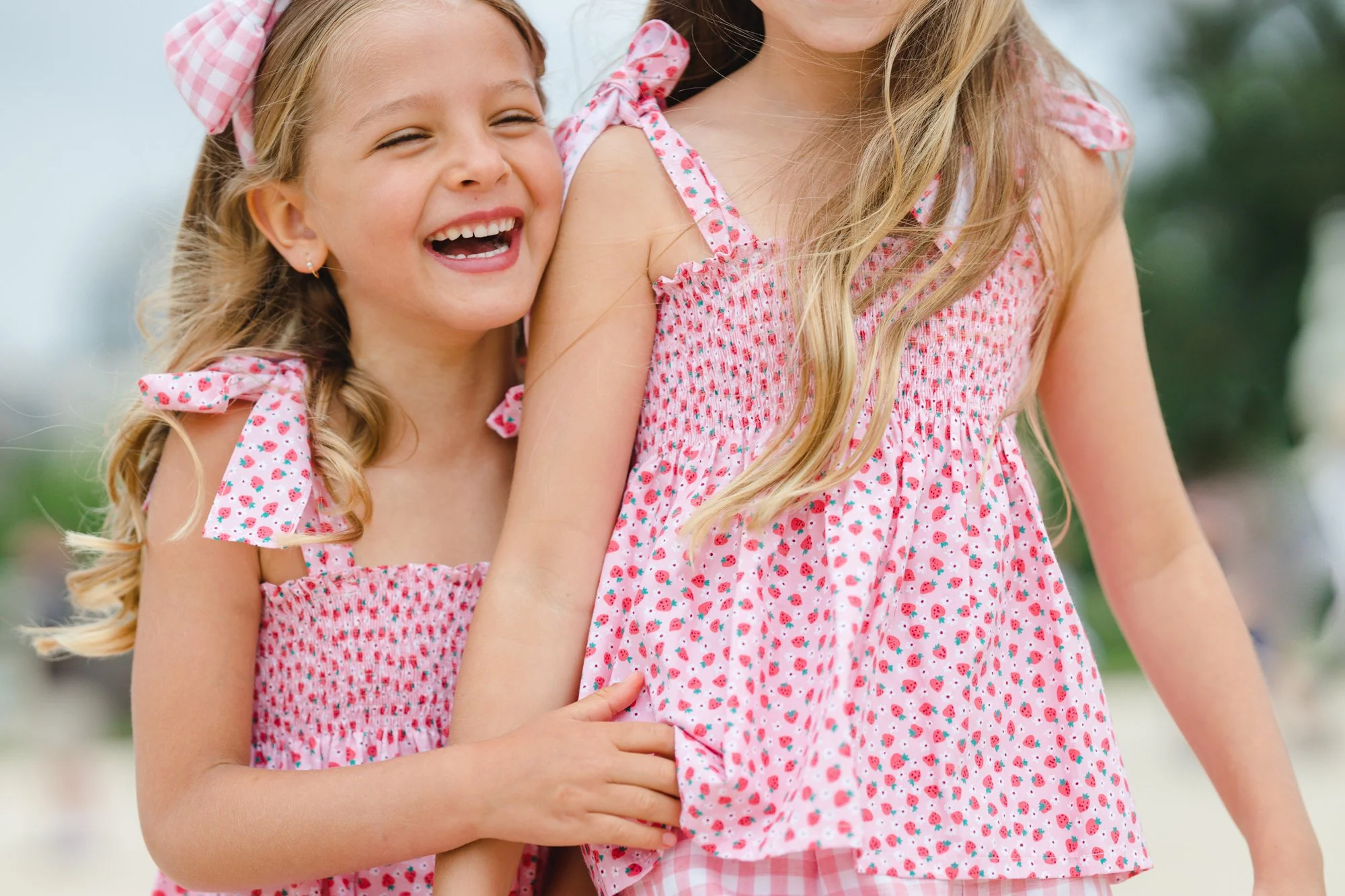 Two young girls wearing matching pink and white strawberry print dresses laughing and playing together outdoors.