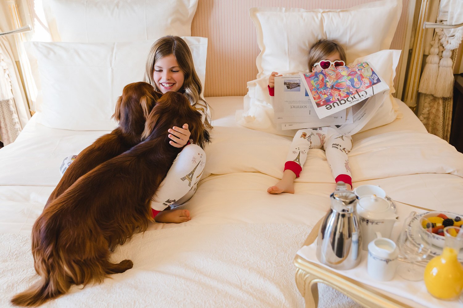 Two young girls relaxing on a bed, one with a newspaper and wearing sunglasses, the other petting two brown dogs, all in a cozy Ritz Paris bedroom.