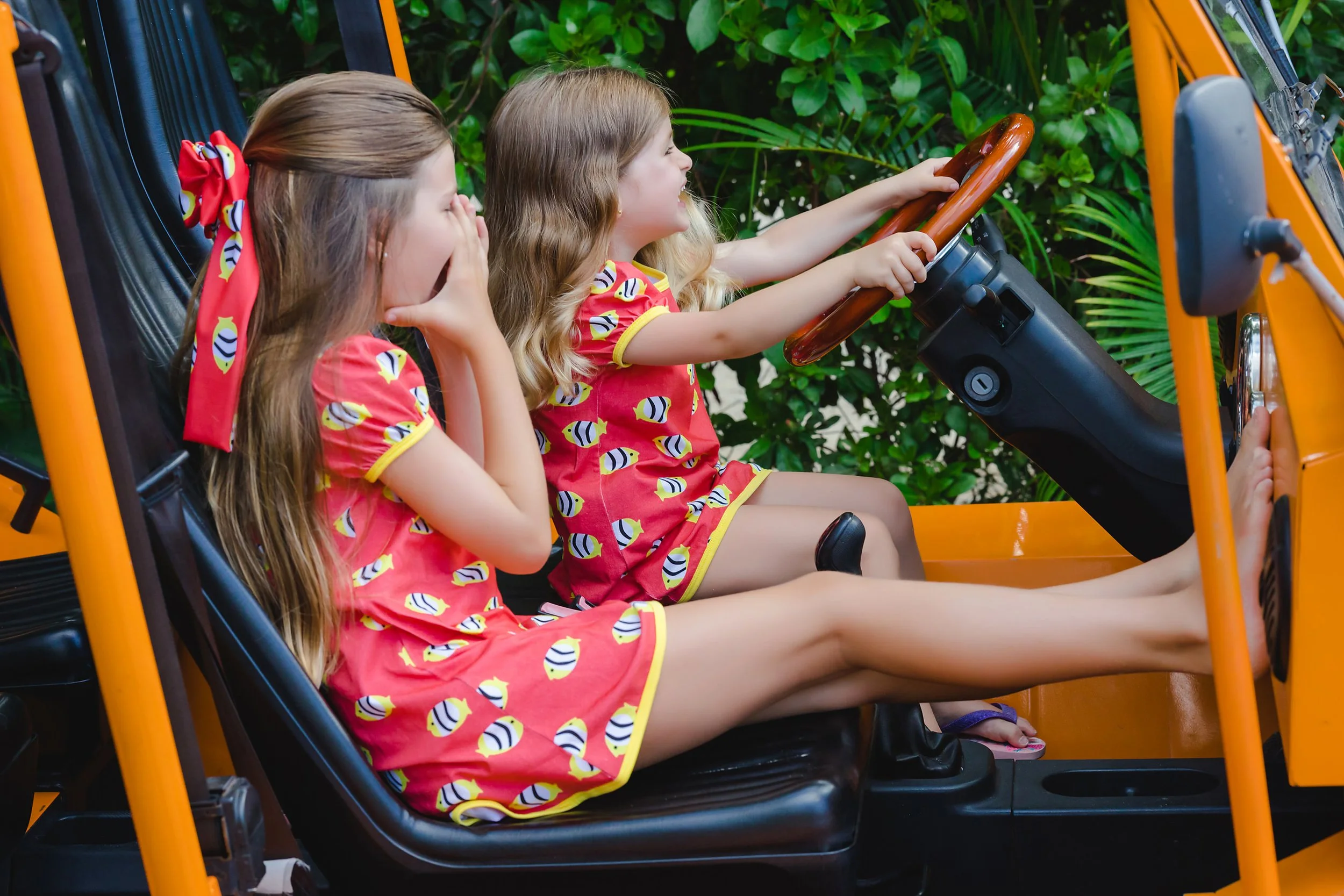 Two young girls in matching red and yellow bee-themed dresses riding in a yellow off-road vehicle, with one girl driving and the other covering her mouth in surprise, surrounded by green foliage.