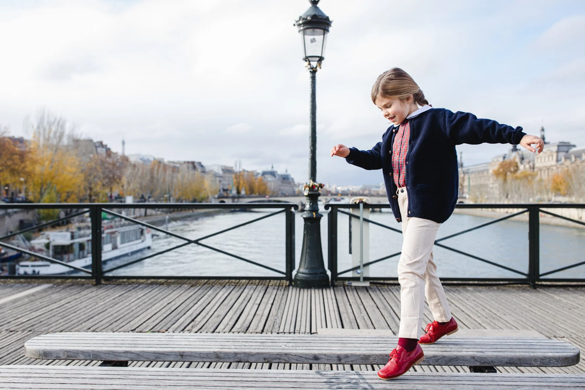 A young girl balancing on a wooden bench on a bridge over a river in Paris. She is wearing a navy blue jacket, a red plaid shirt, cream pants, and red shoes. Trees with autumn leaves line the riverbank, and buildings are visible in the background.