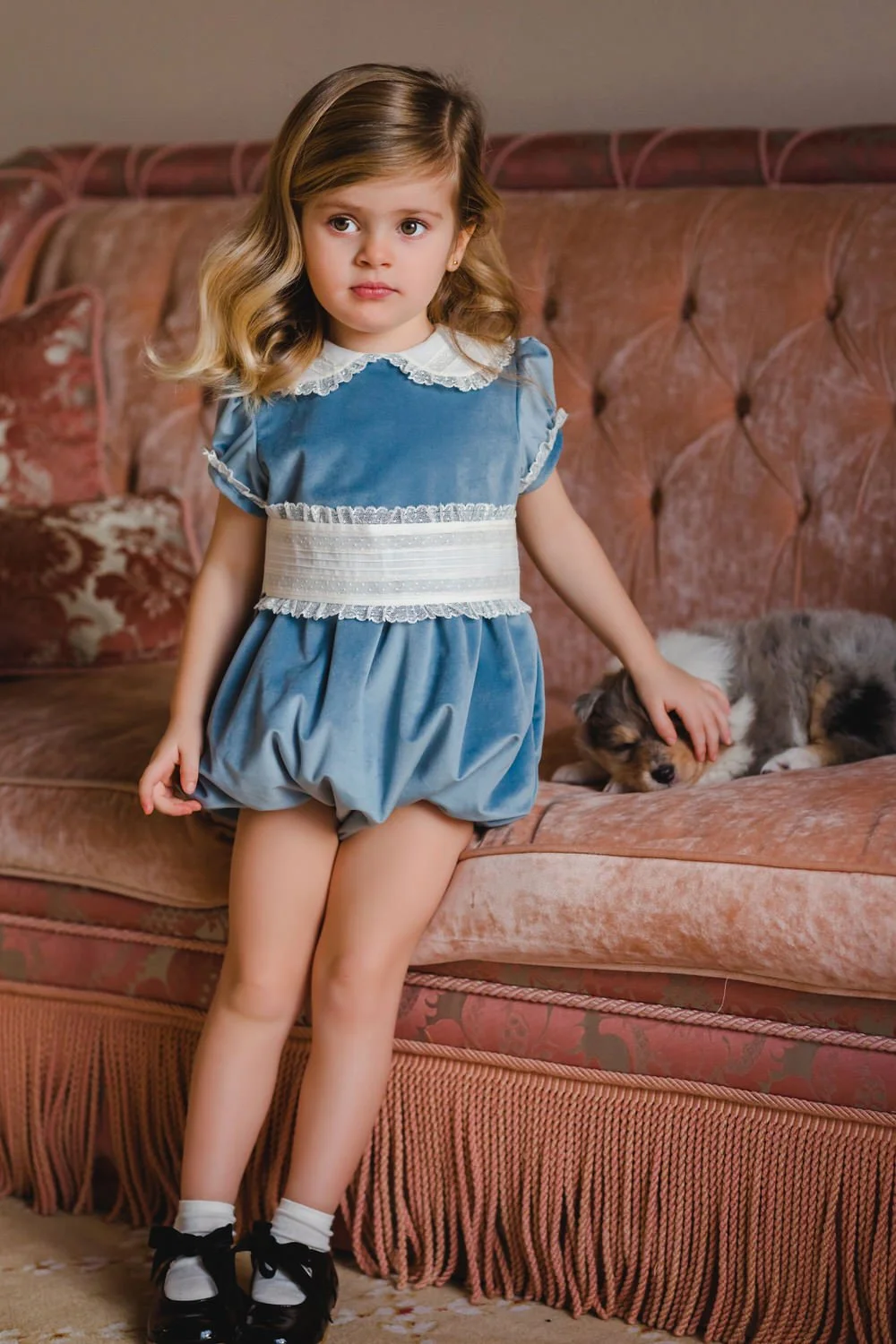 A young girl with wavy blonde hair, wearing a blue velvet dress with lace details, standing on a vintage pink sofa, petting a sleepy puppy.