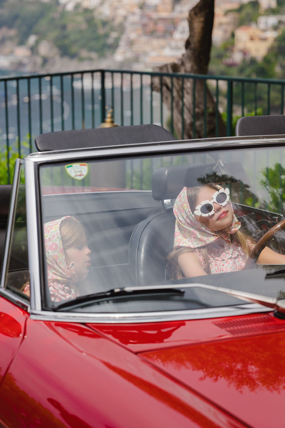 Two young women in vintage clothing and wrap-around sunglasses sitting in a red convertible car. One woman is driving, and the other is passenger. They are outdoors in Positano,  a tree in the background.