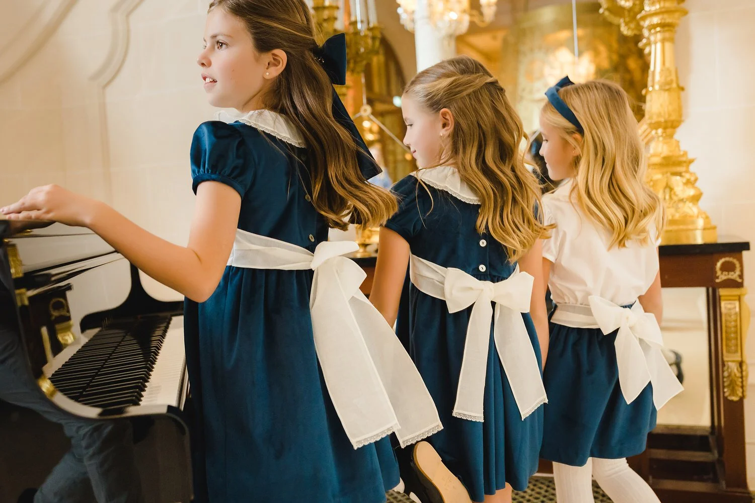 Four young girls in matching Blue dresses and white aprons with large bows, standing beside a piano in an elegant room, possibly participating in a choir or recital.