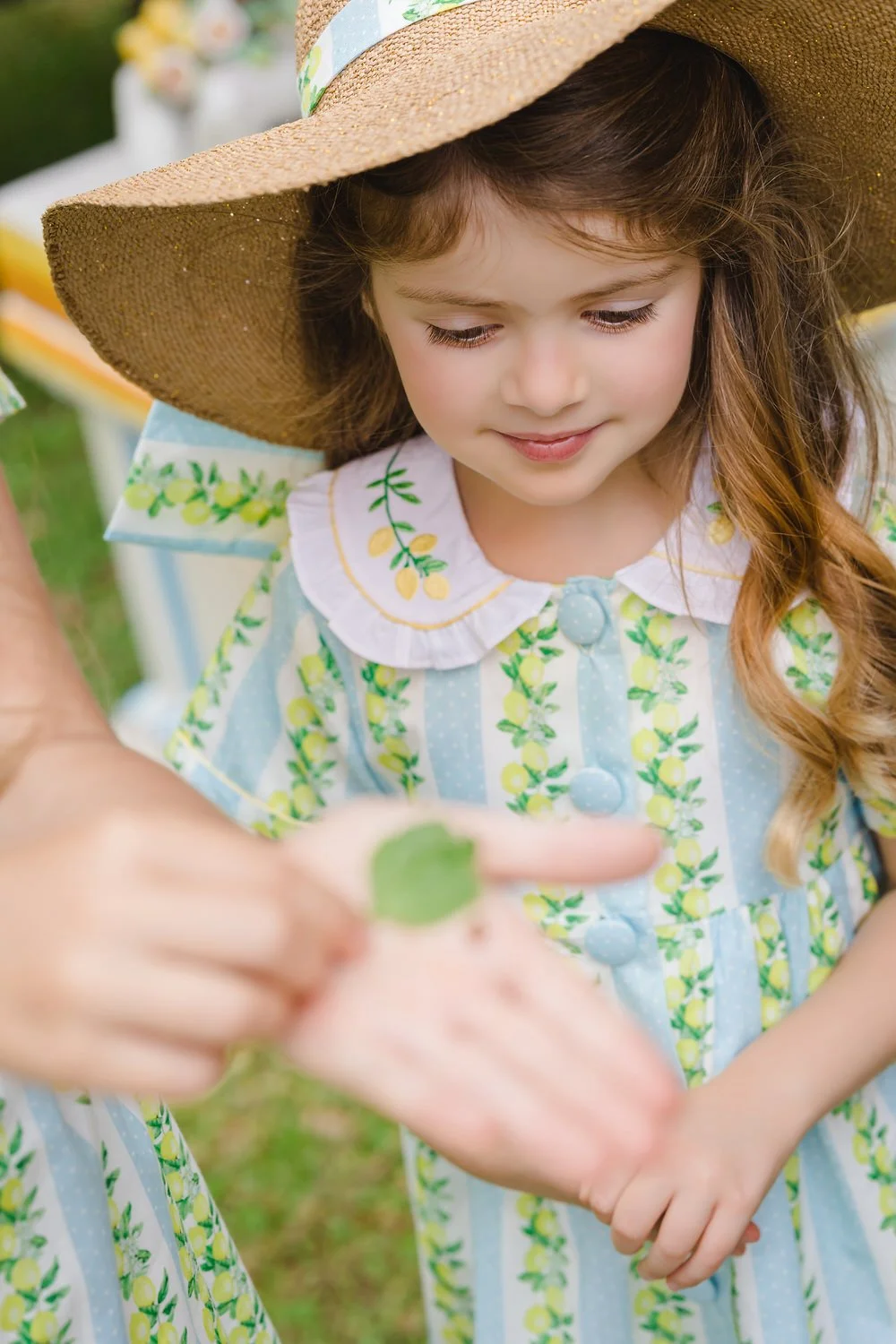 A young girl with long brown hair wearing a sun hat and a dress with lemon and green fruit print, looking at a hand holding a small leaf during an outdoor activity.