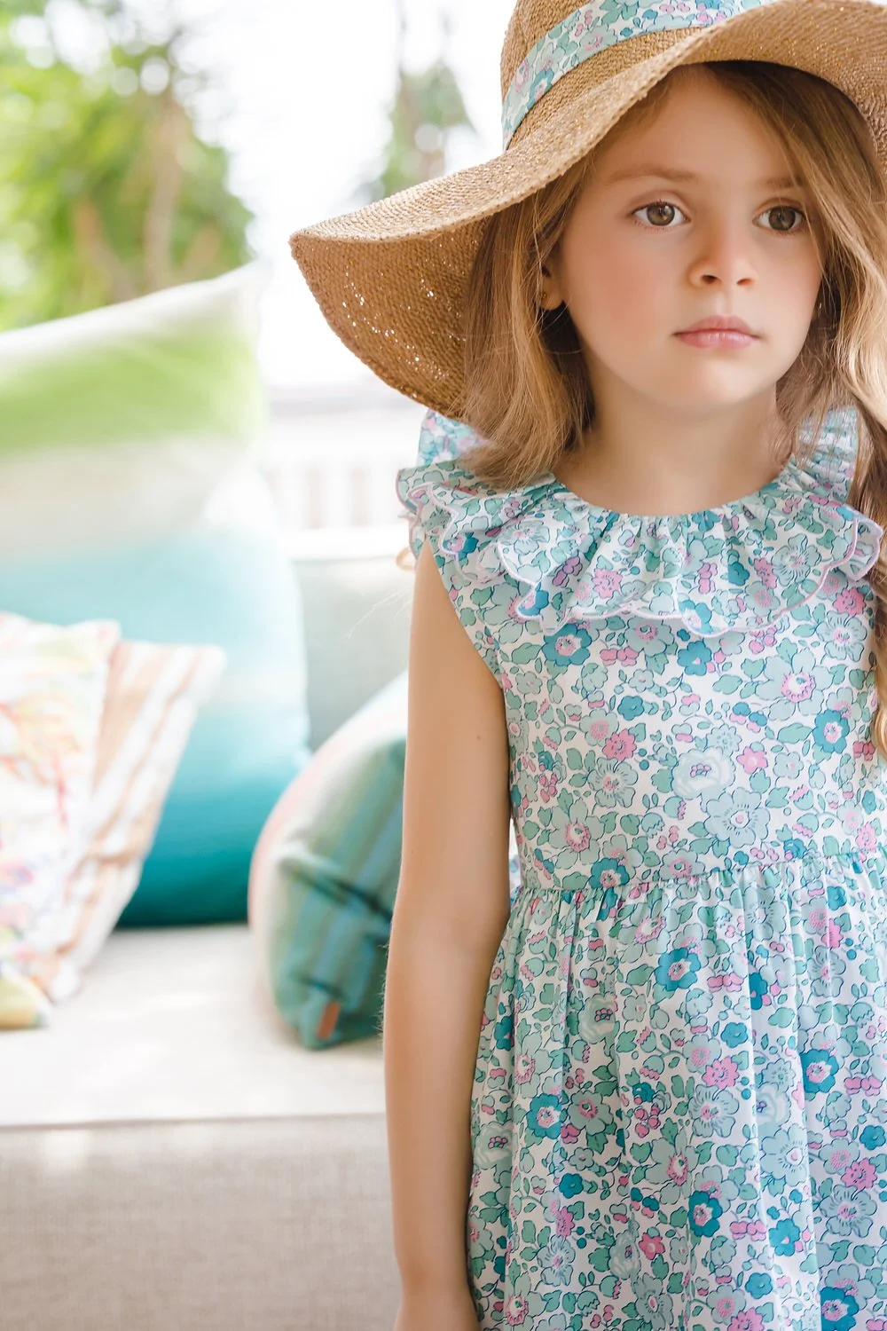 A young girl wearing a floral dress and a large straw hat standing indoors with a sofa and cushions in the background.