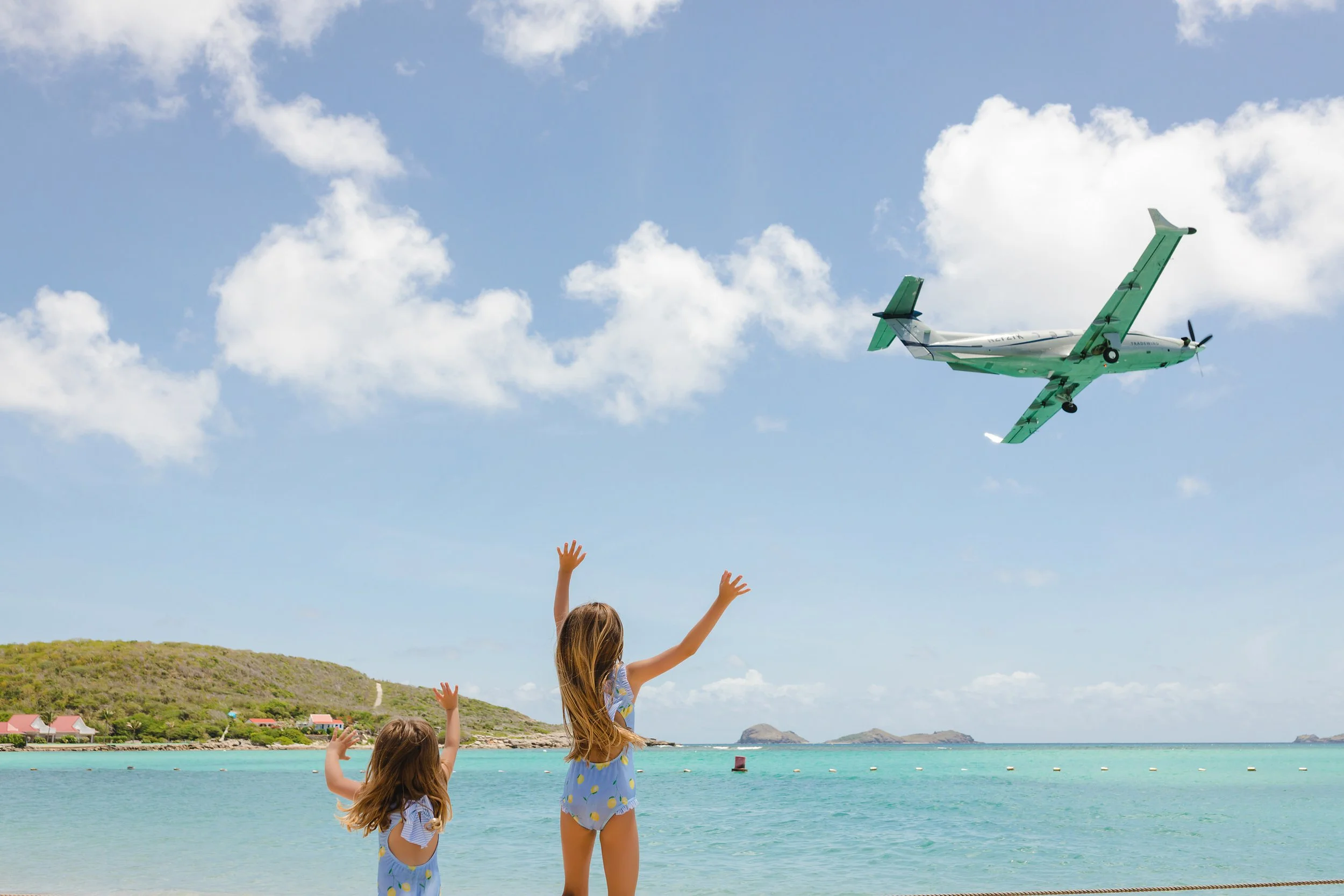 Two young girls in matching blue swimsuits with yellow polka dots waving at a seaplane flying overhead on a clear, sunny day at the beach.