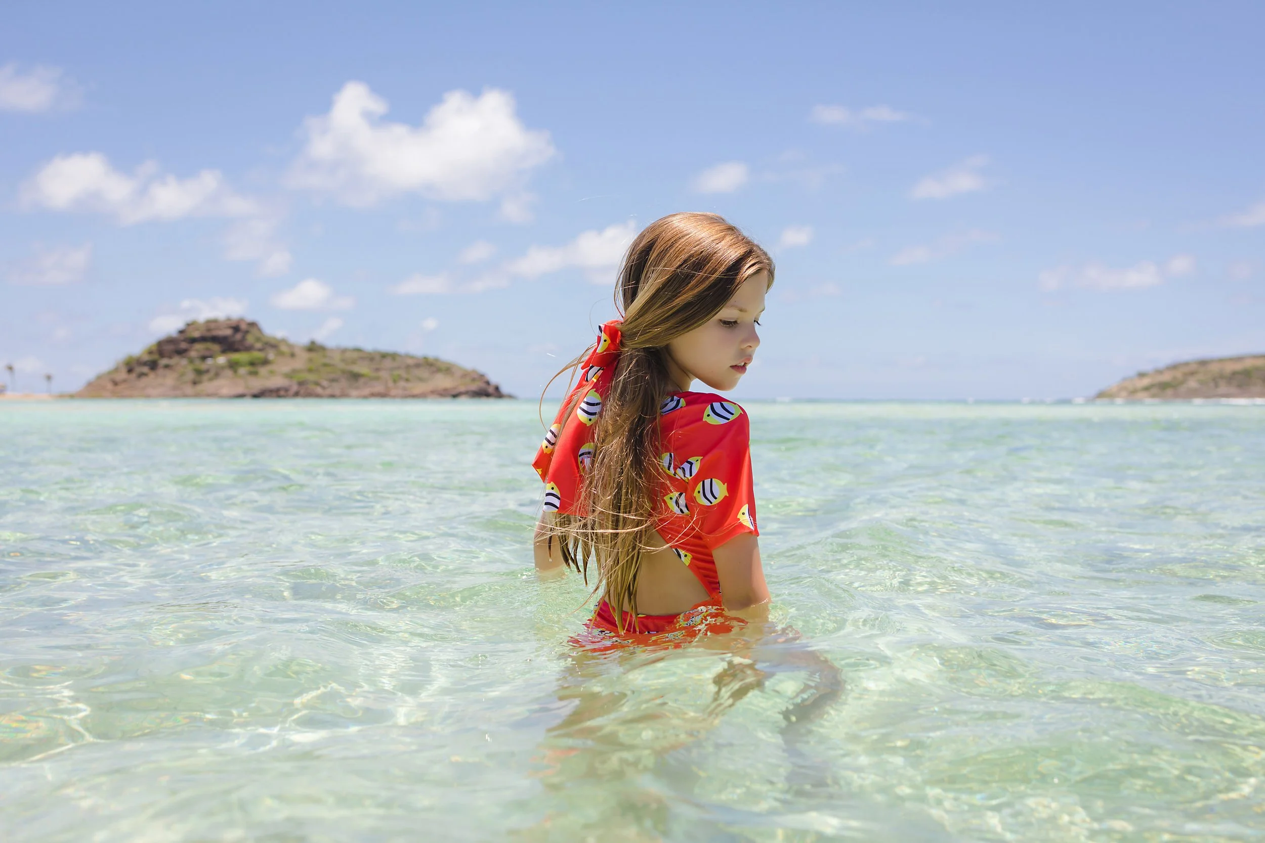 A young girl with long hair wearing a red swimsuit with a colorful pattern stands in shallow clear water at the beach, with small islands in the background on a sunny day.