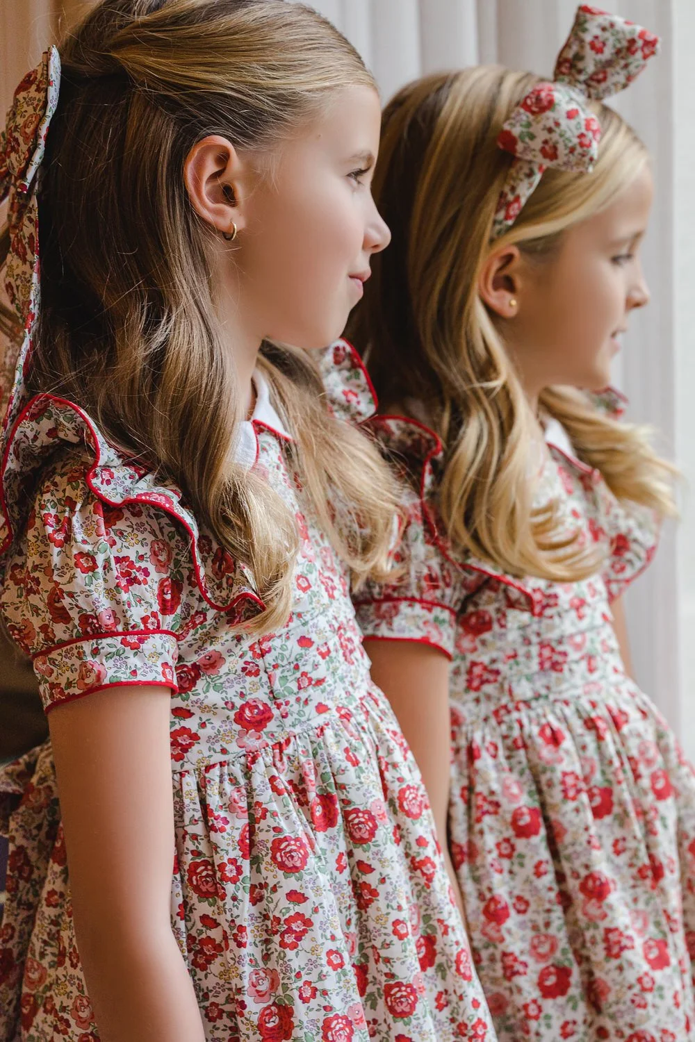 Two young girls wearing matching vintage floral dresses with bows in their hair, standing side by side and looking out a window.