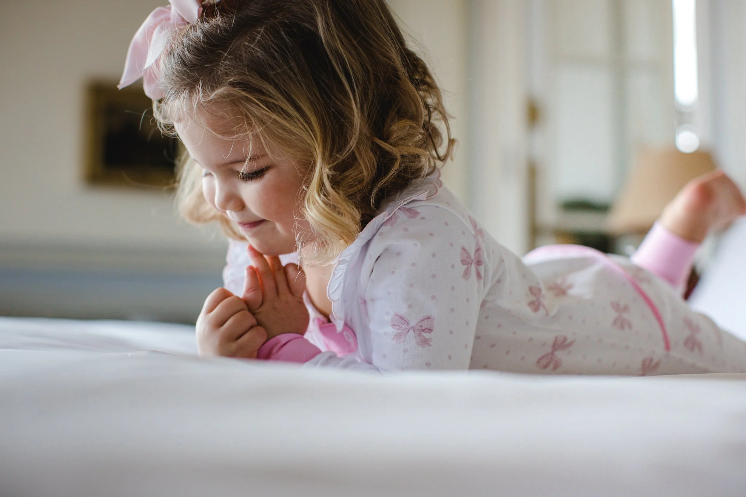 A young girl with blonde curly hair and a pink bow headband lying on her stomach on a bed, clasping her hands together and smiling.