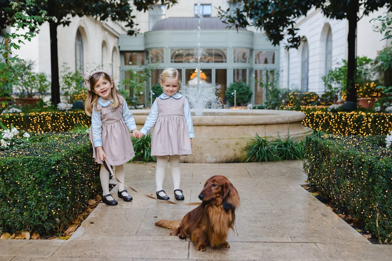 Two young girls in beige dresses and white tights holding hands, standing on a pathway in front of a large fountain, in the garden of Ritz Paris, with a golden retriever dog sitting nearby, surrounded by festive holiday lights and greenery.