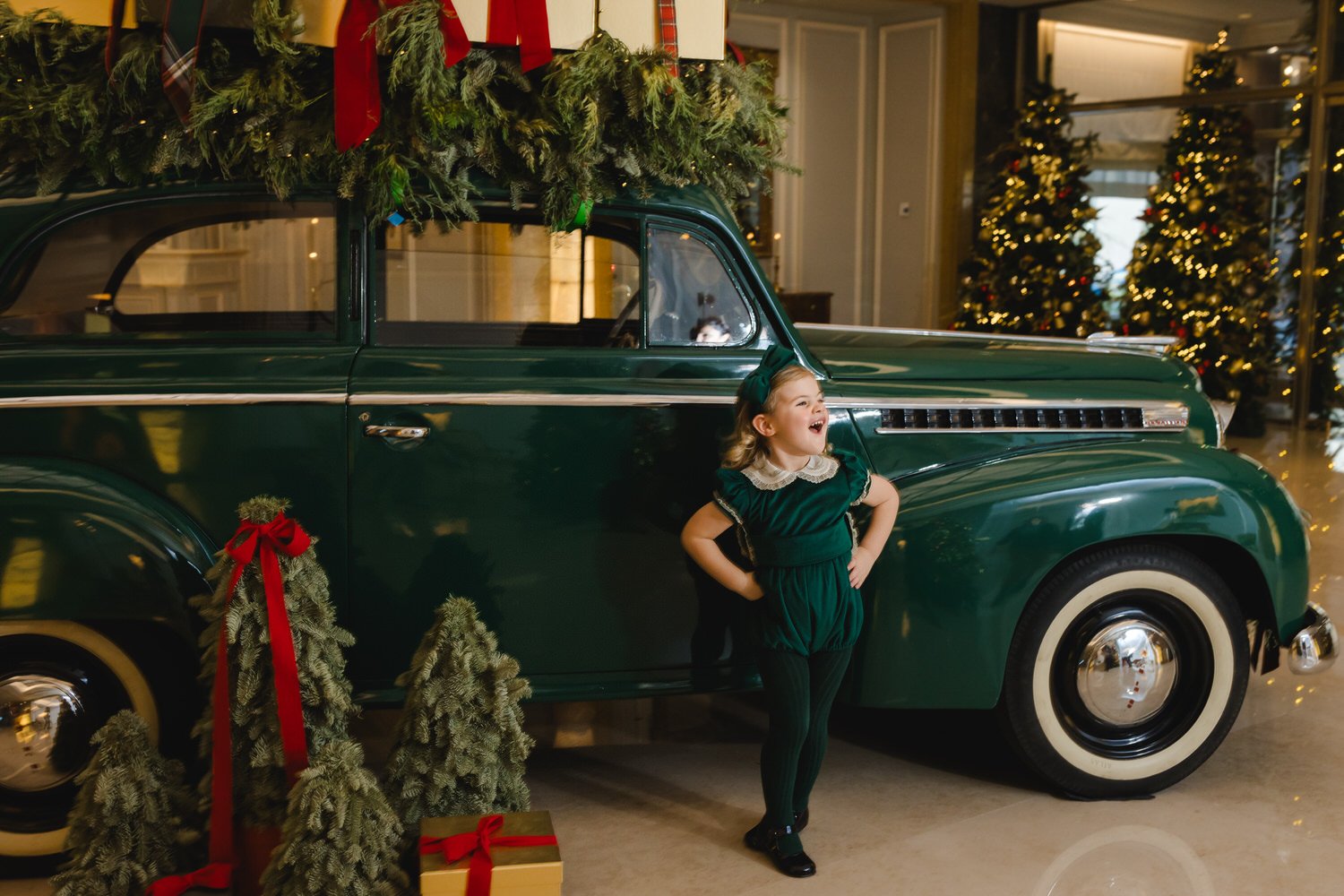 A young girl in a dark green dress with a white collar and a matching headband is standing next to a vintage green car decorated with Christmas greenery and red ribbons. In the background, there are decorated Christmas trees with lights, and wrapped 
