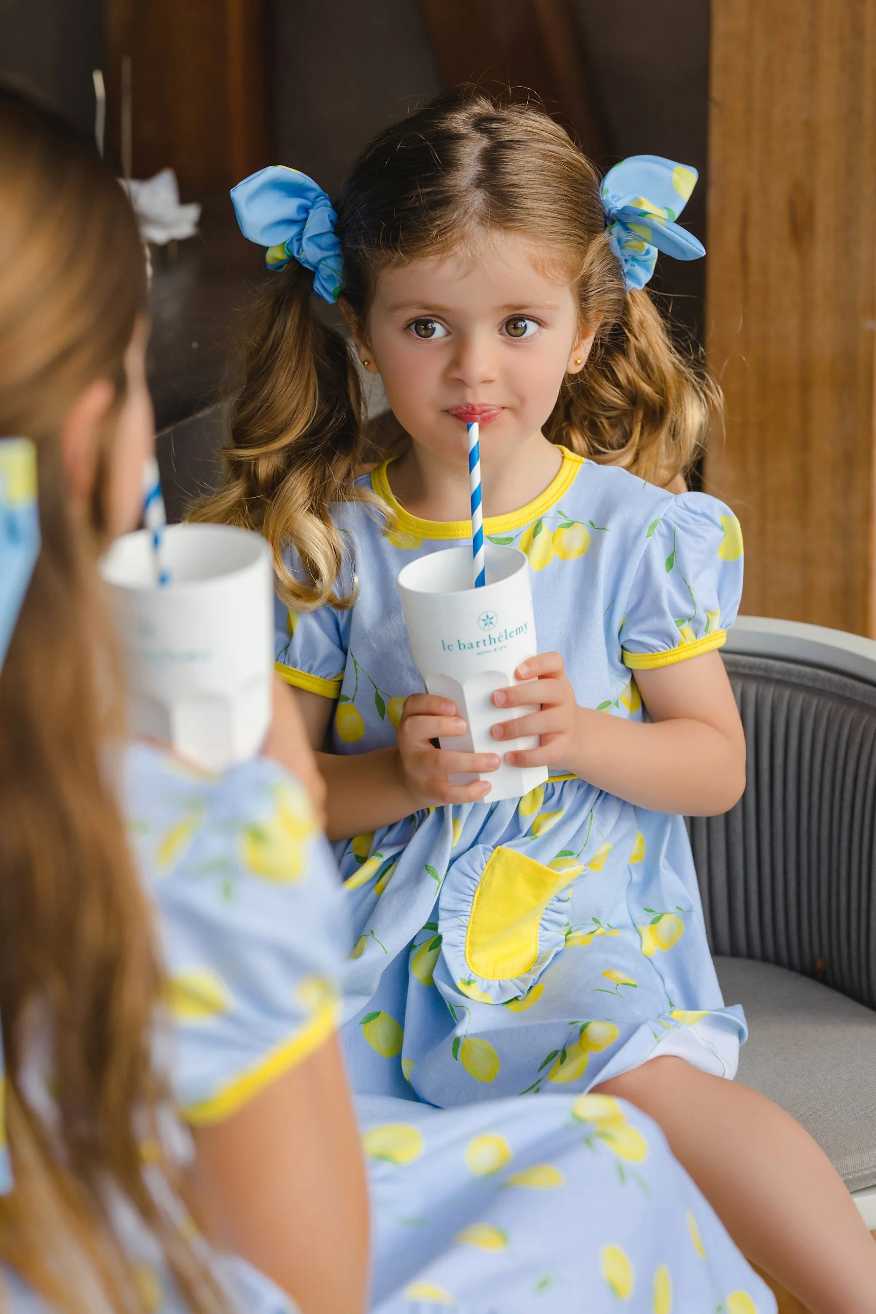 A young girl with light brown curly hair in pigtails and blue bows, wearing a light blue dress with lemon print, sipping from a white cup with a blue and white striped straw, sitting on a bench indoors.