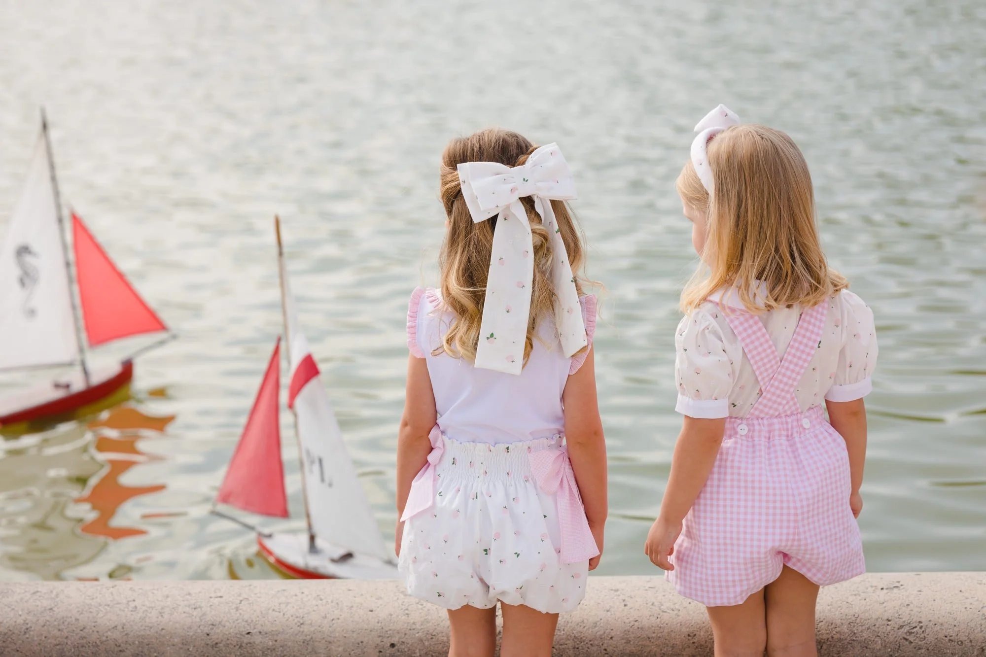 Two young girls with bows in their hair standing by a body of water, watching sailboats with red and white sails.