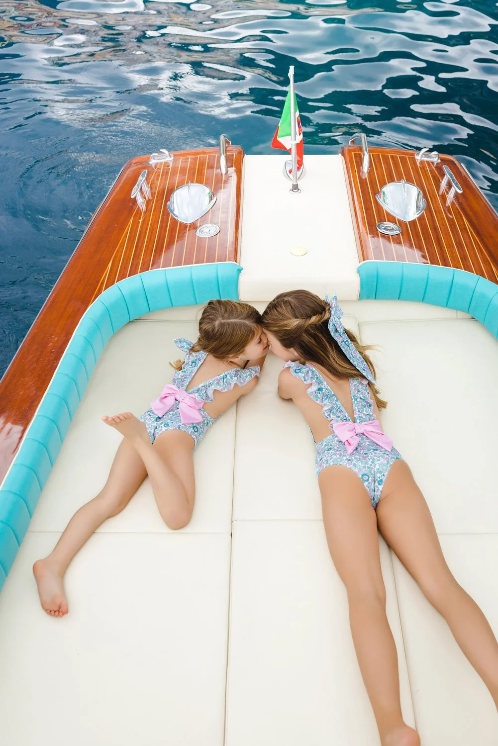 Two young girls in matching swimsuits with pink bows are lying on a boat Riva in Positano, kissing each other with water in the background.
