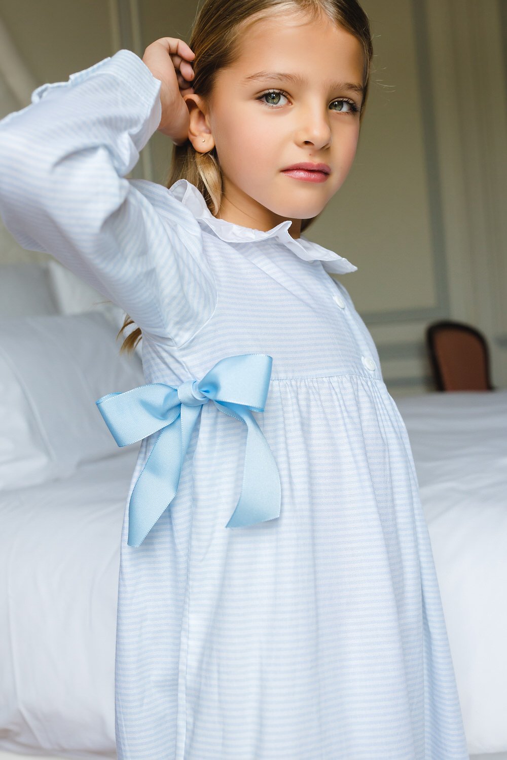 Young girl in a white dress with a light blue bow, standing in a room with a bed in the background, touching her hair.