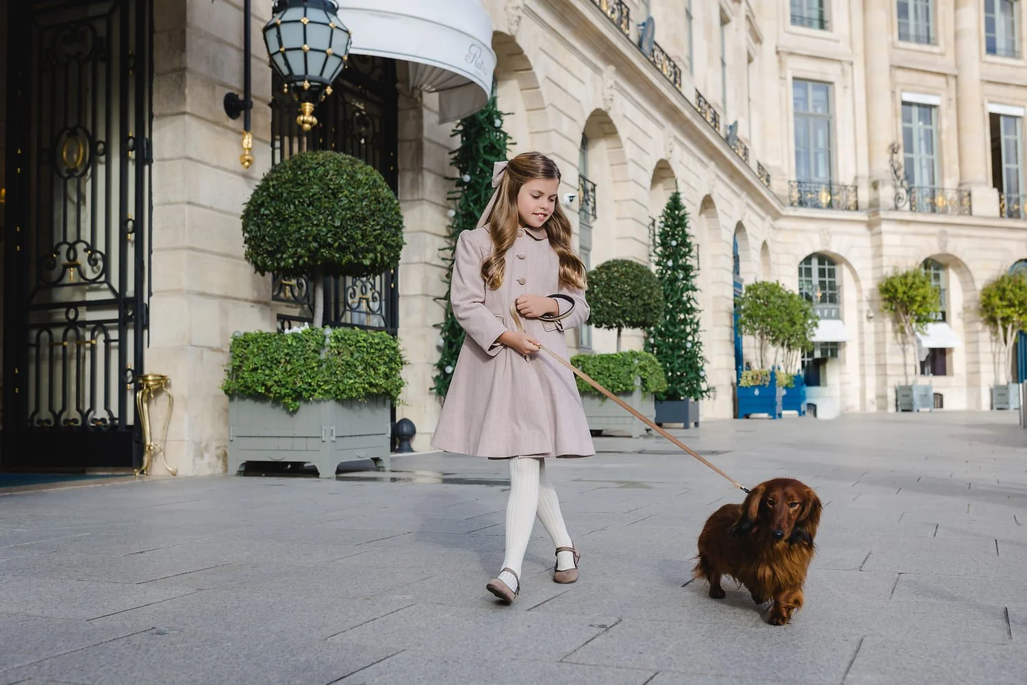 A young girl in a beige coat and white tights walking a small brown dachshund on a leash outside the luxurious Ritz Paris with potted plants and decorative exterior.