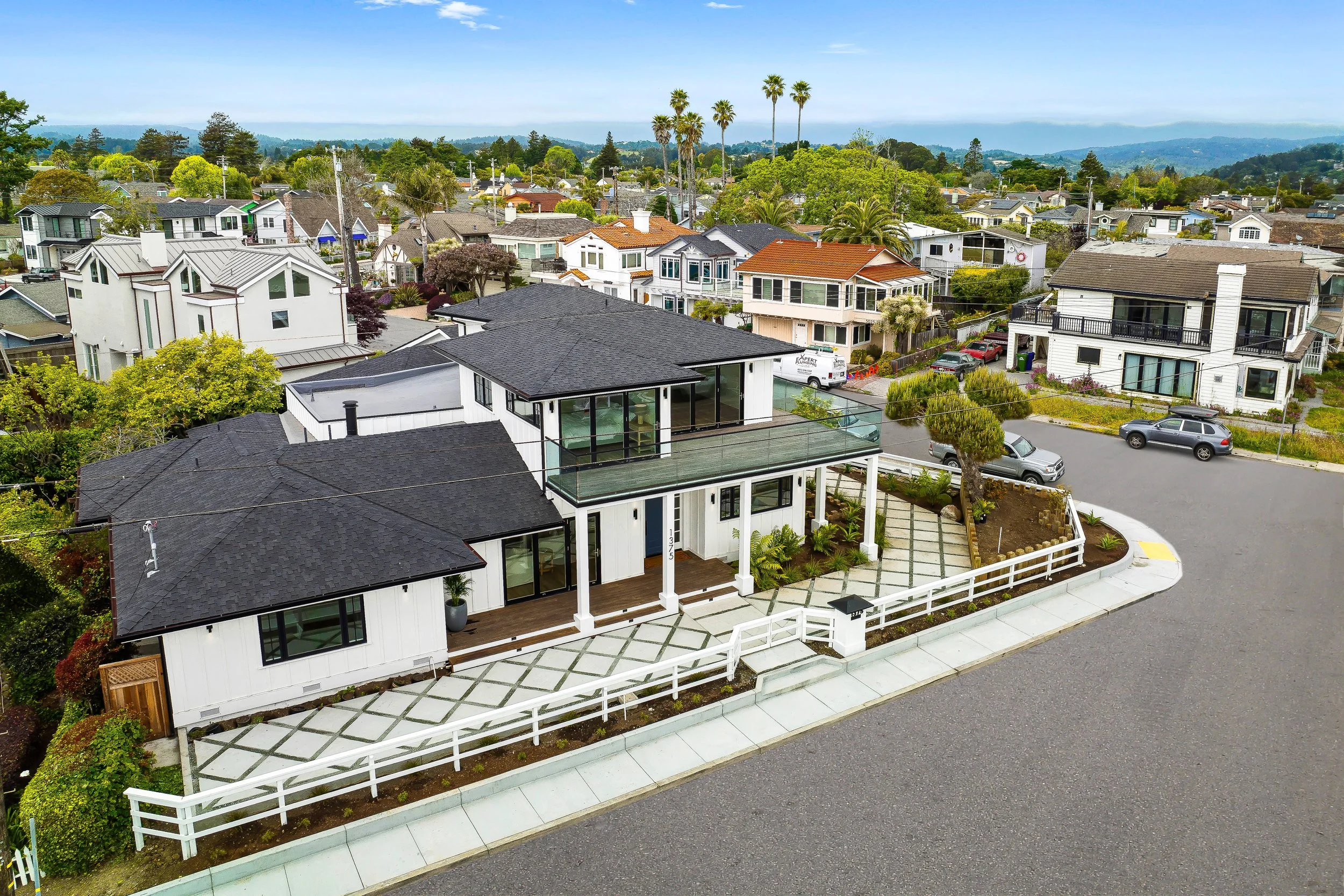 A modern, two-story white house with black roof and large glass balcony in a suburban neighborhood, surrounded by other houses, trees, and parked cars in Capitola, California.