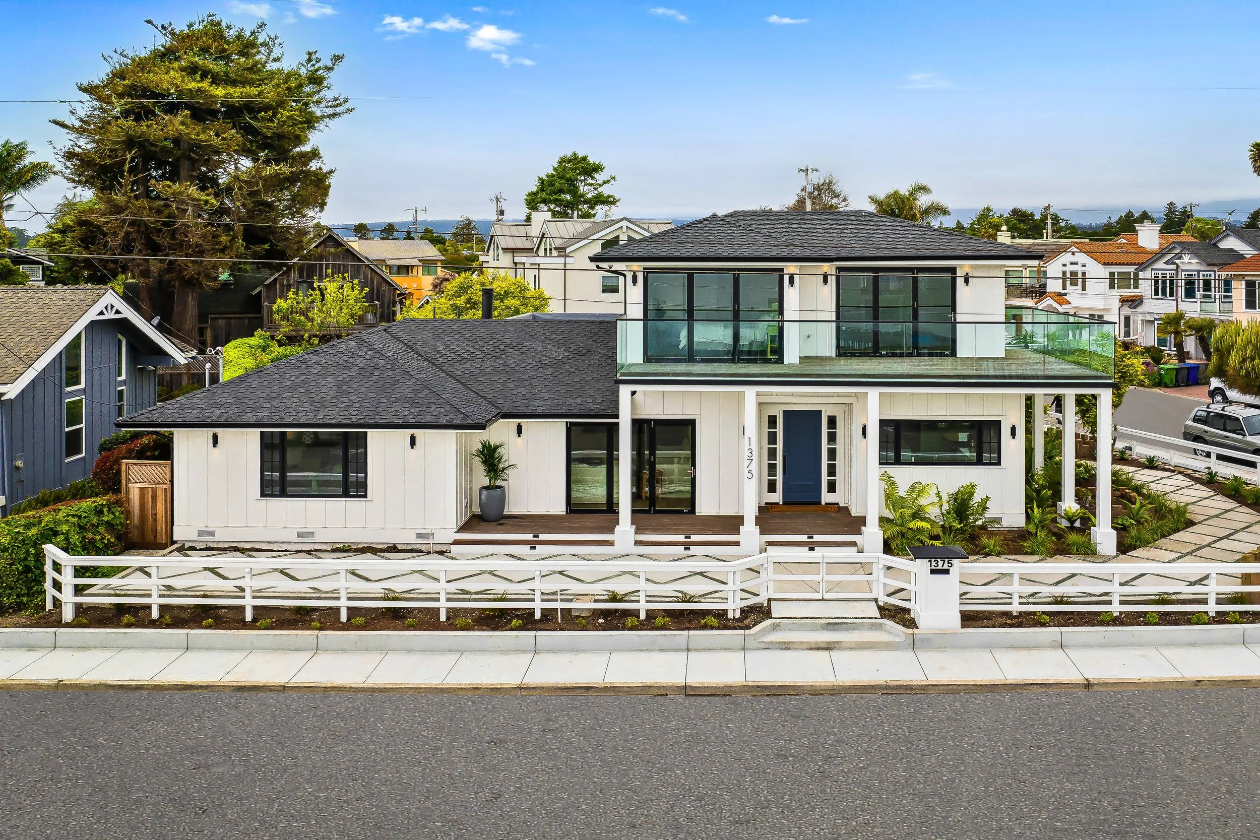Modern white two-story house with black roof, glass balcony, wooden deck, and front yard with plants, located in a neighborhood with other houses and trees.