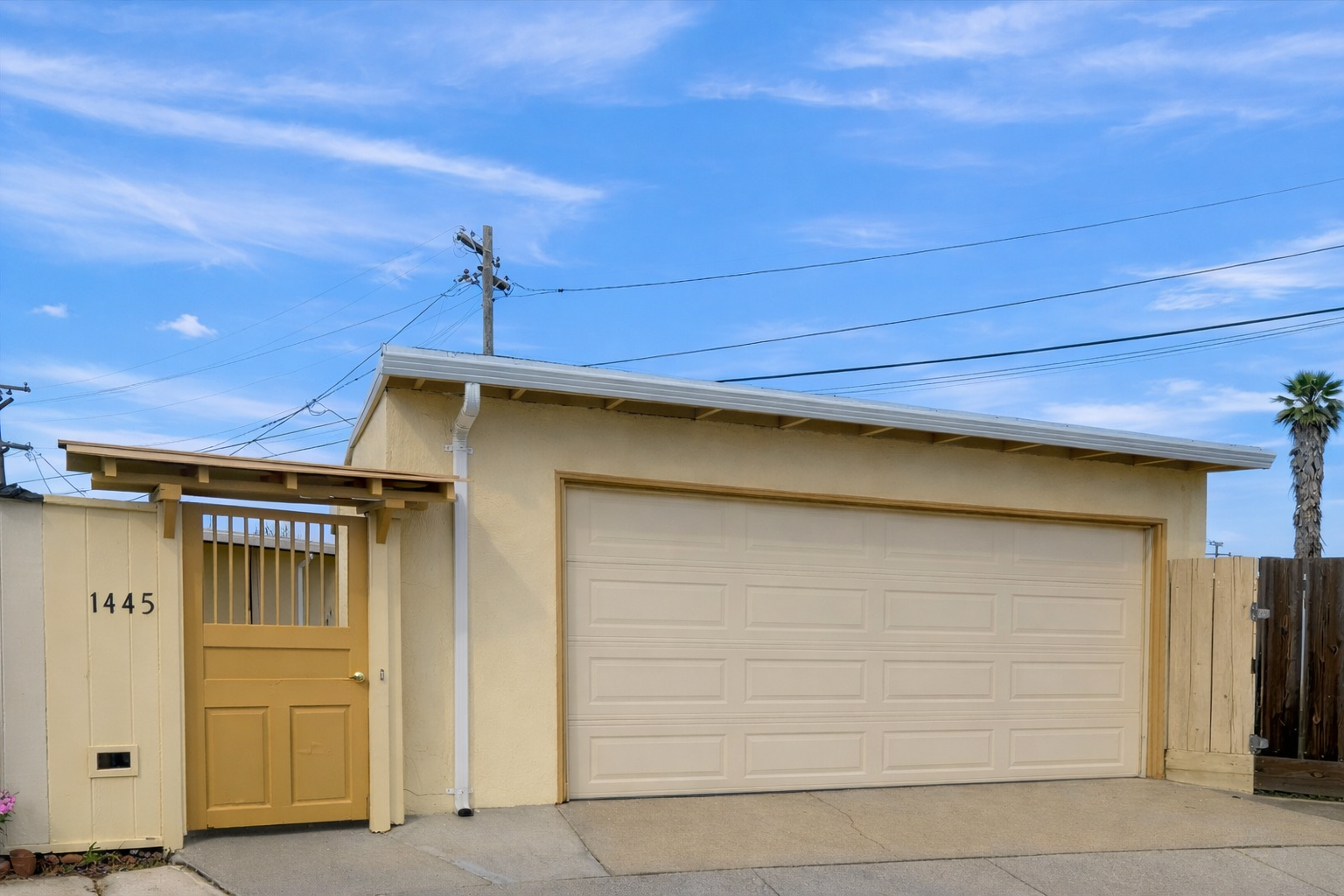 A beige detached garage with a large garage door and a small side gate. The house number 1445 is on the wall beside the side gate. The sky is partly cloudy with blue sky and some thin clouds, and there are electrical poles and lines in the background