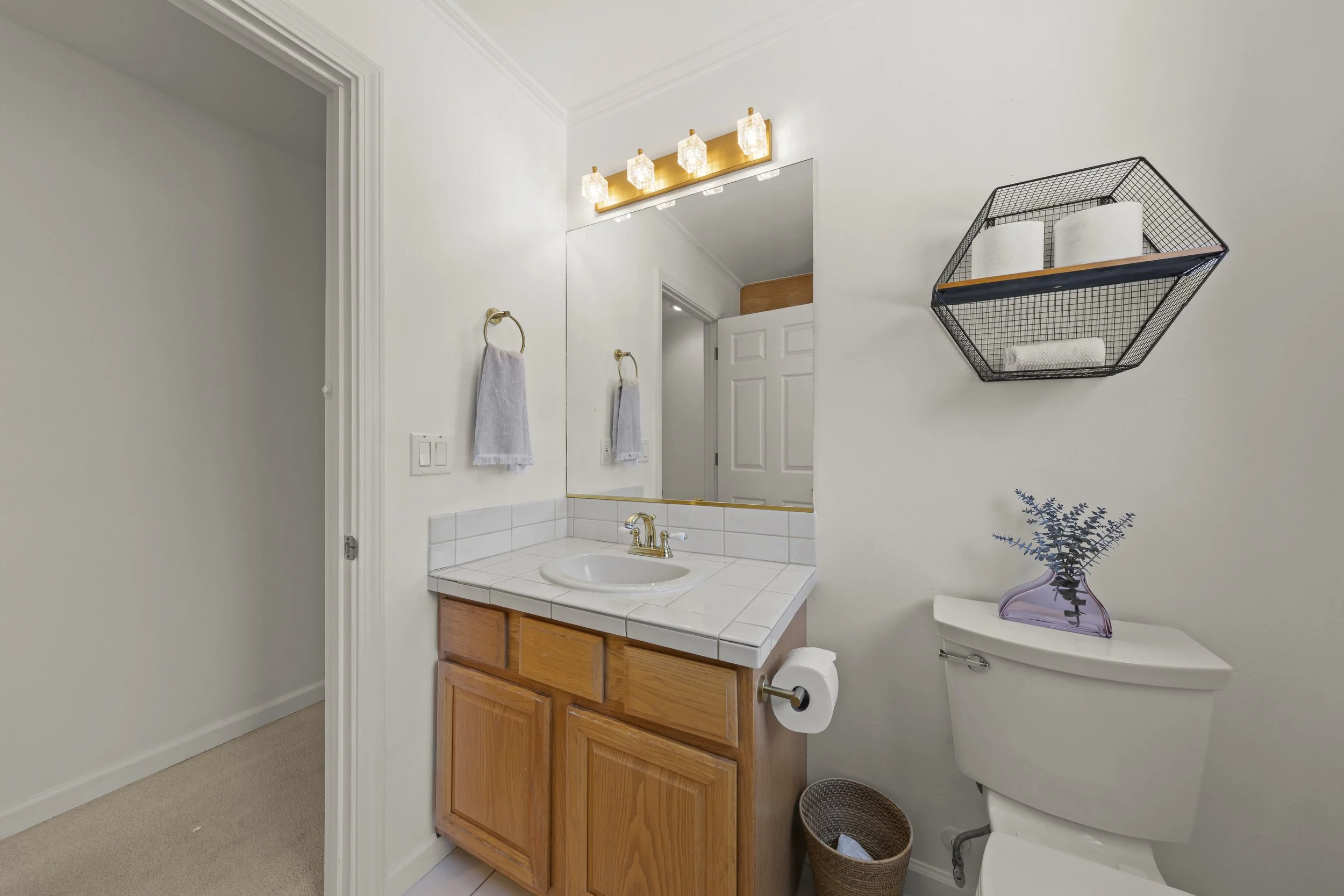 Small bathroom with a white tiled vanity, wooden cabinet, large mirror, wall-mounted light fixture, gray towels on gold rings, black metal wall shelf with toilet paper rolls, and a decorative purple vase with purple flowers on the toilet tank.