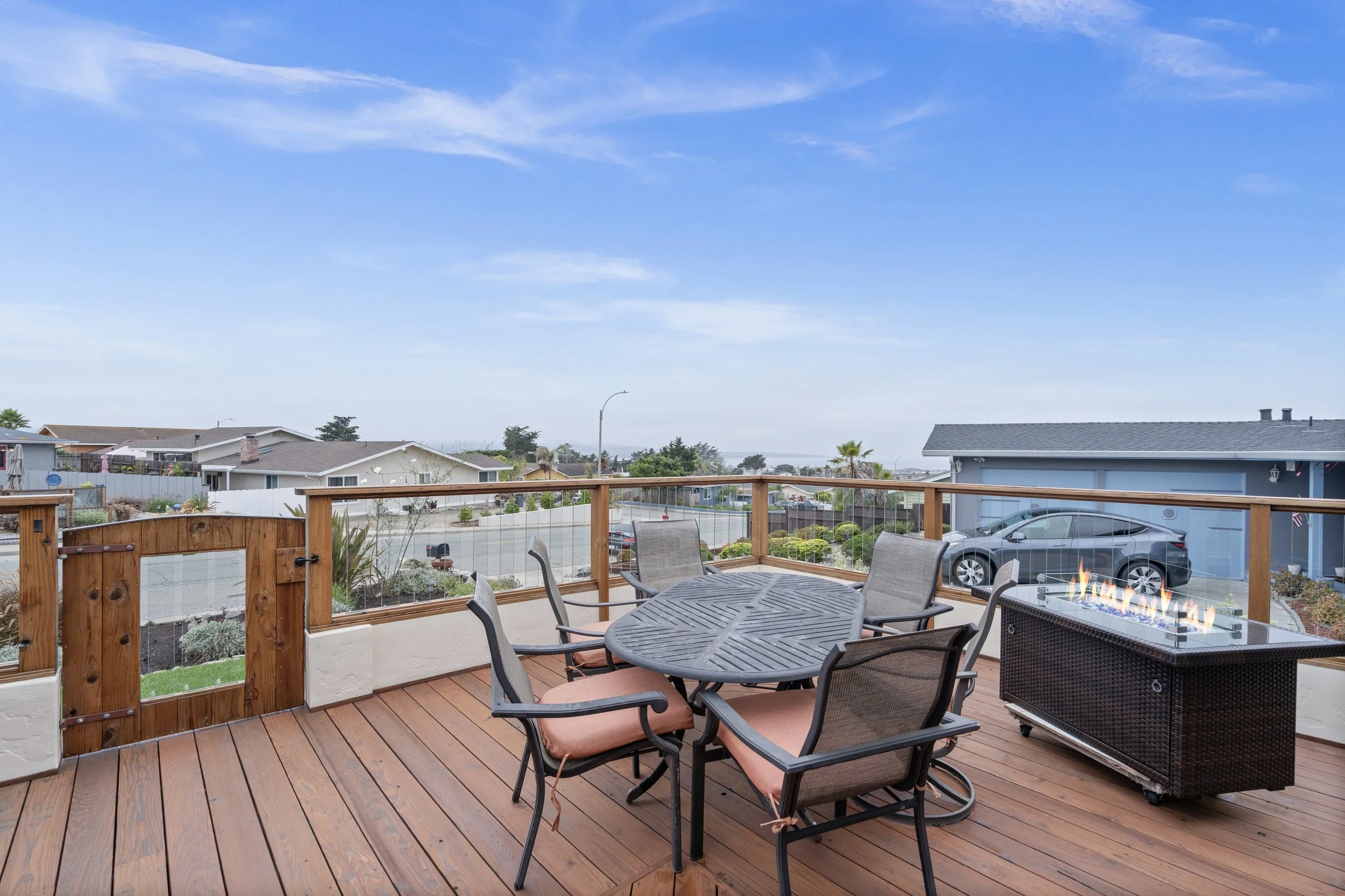 A wooden deck with outdoor furniture and a fire pit, overlooking a suburban neighborhood under a partly cloudy sky.