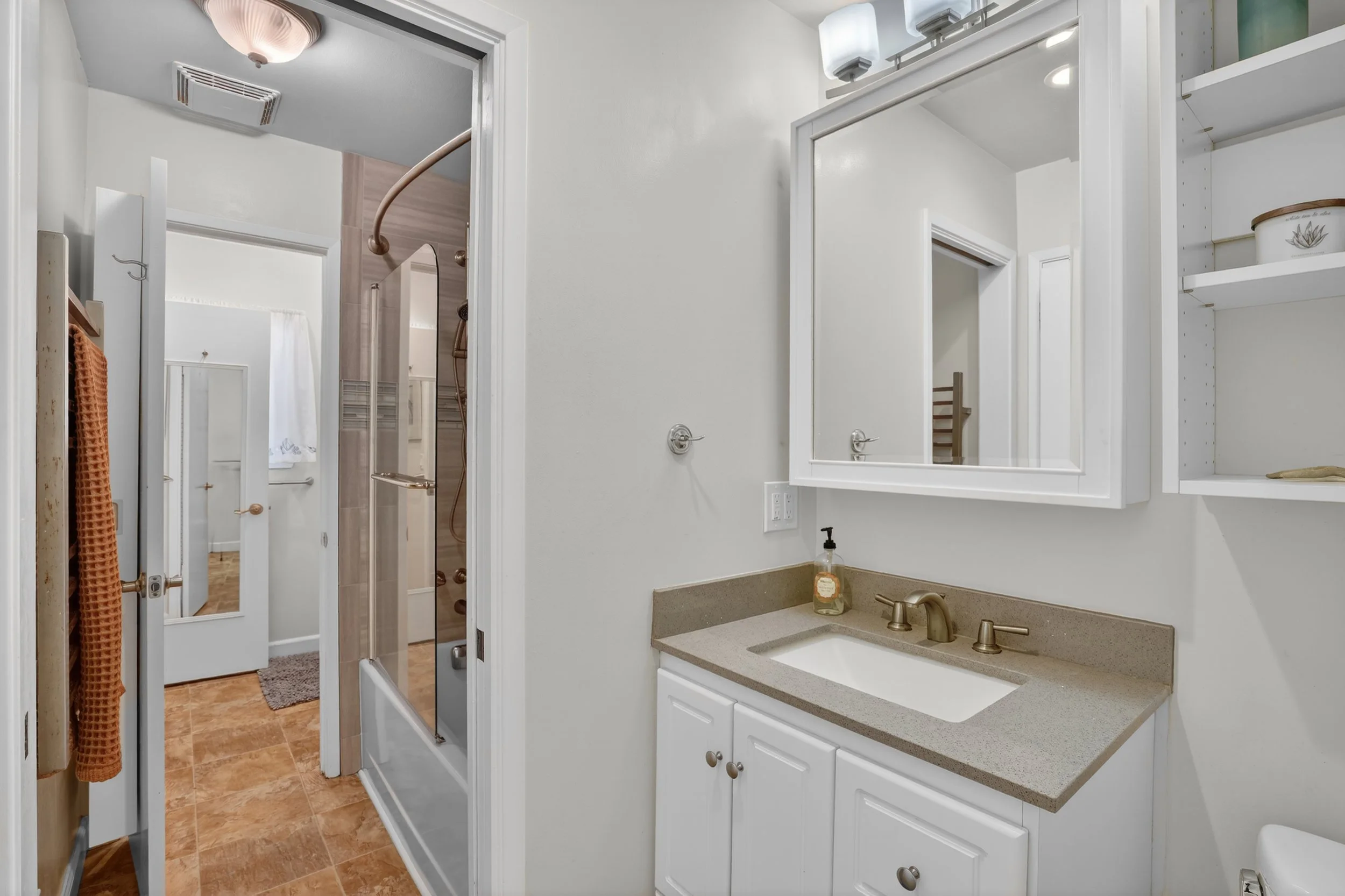 Bathroom with white walls, a beige countertop vanity with sink, a large mirror, a small shelf with decorative items, visible shower with glass door, and a doorway leading to another room with a mirror and window.