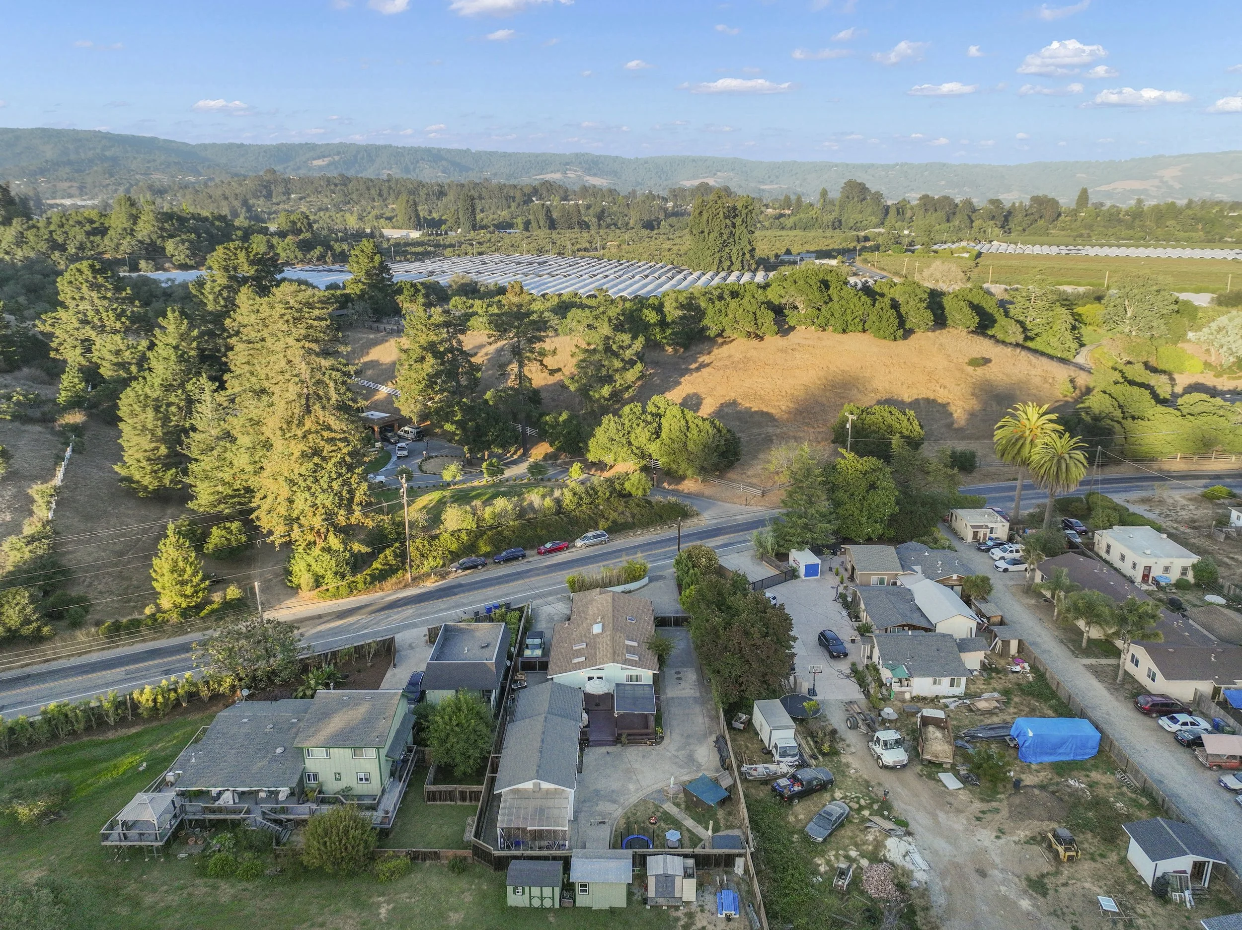 Aerial view of a residential neighborhood with houses, parked cars, and a road, surrounded by trees and hills in the distance.