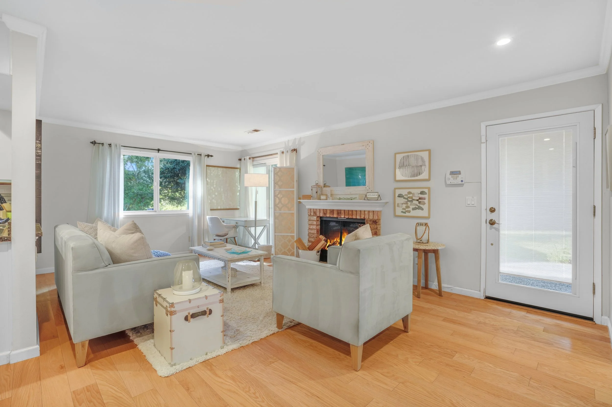 Bright living room with white walls and hardwood floors, featuring a fireplace with a mirror above, a white sofa and armchair, a coffee table, large windows with white curtains, and a door with blinds leading outside.