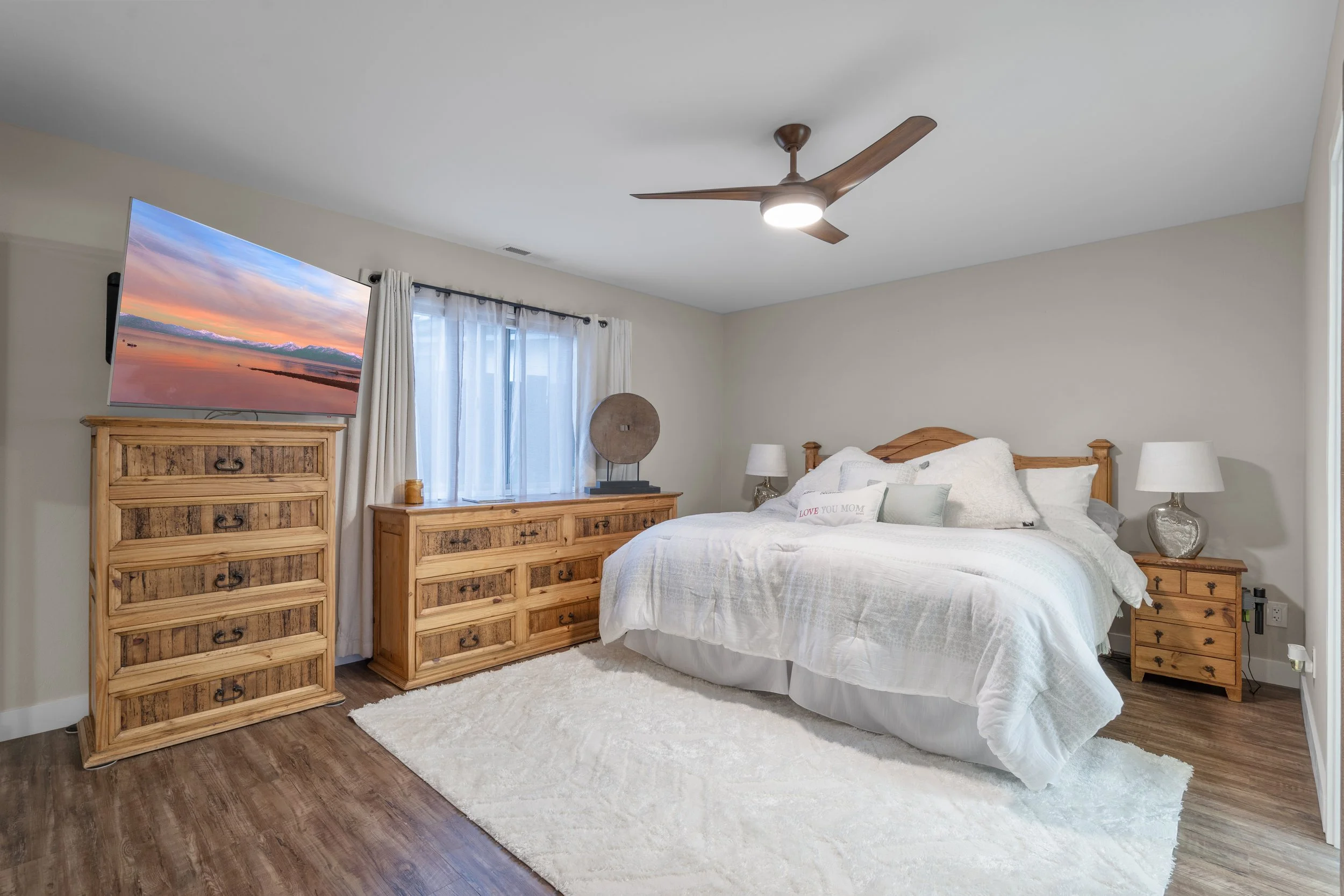 Bedroom with a bed, bedside tables, a wooden dresser, a large TV, and a window with curtains.