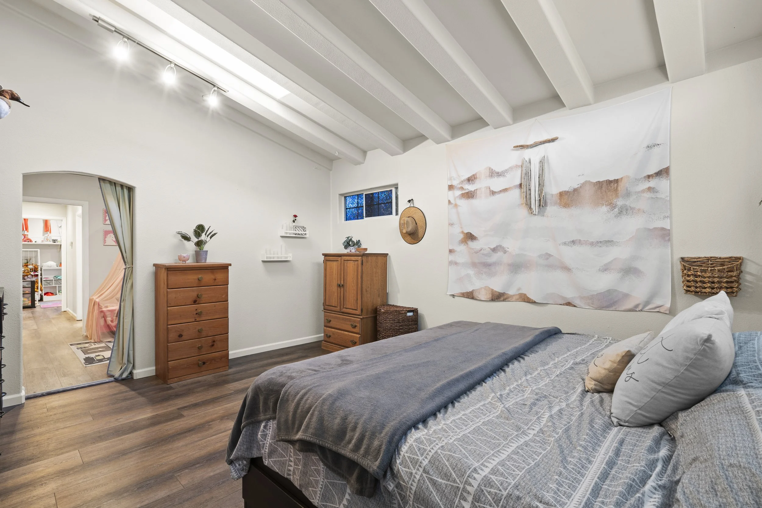 A cozy bedroom with a bed covered in a patterned gray blanket and pillows, wooden dressers, a wall hanging, small wall shelves, a window, and a doorway leading to a playroom with pink and orange decor.
