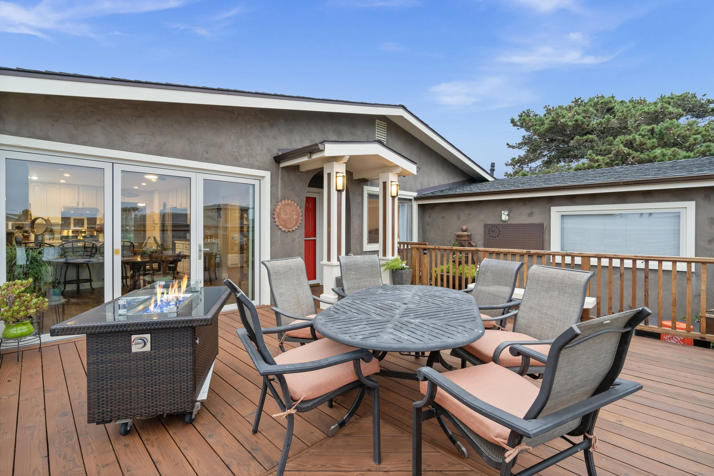 Outdoor wooden deck with six chairs around a round table, a gas fire pit, and part of a house with large glass sliding doors and windows, on a clear day.