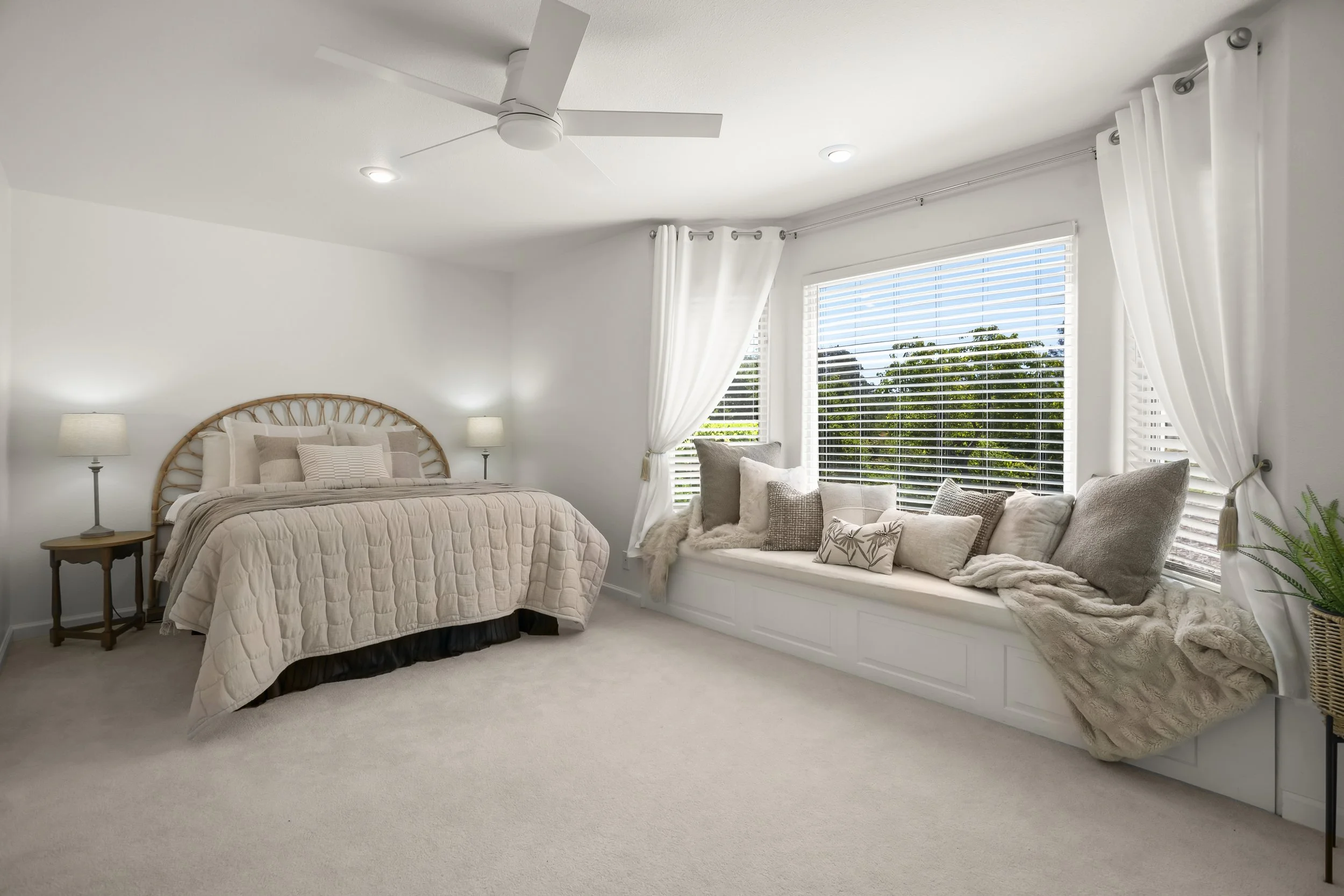 Bright bedroom with a bed on the left, beige quilt, and pillows. Two bedside lamps with lampshades. Large window seat on the right with multiple pillows and a throw blanket. White curtains and blinds, ceiling fan, and potted plant in a woven basket in the corner.