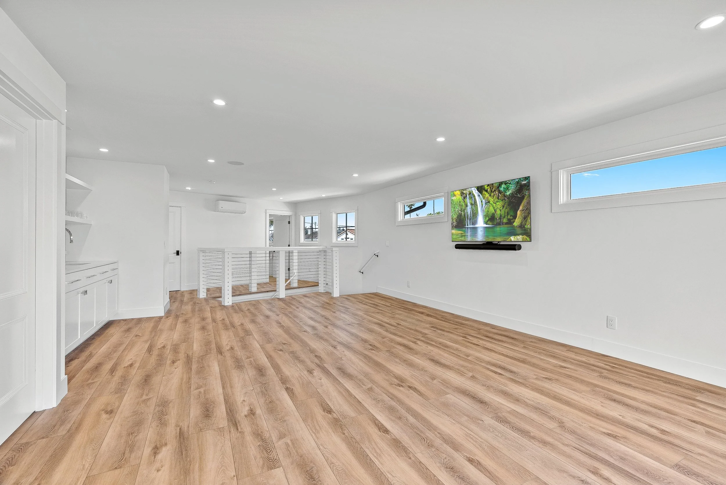 Empty living room with wood flooring, white walls, multiple small windows, a wall-mounted TV, and a white railing in Capitola, California.