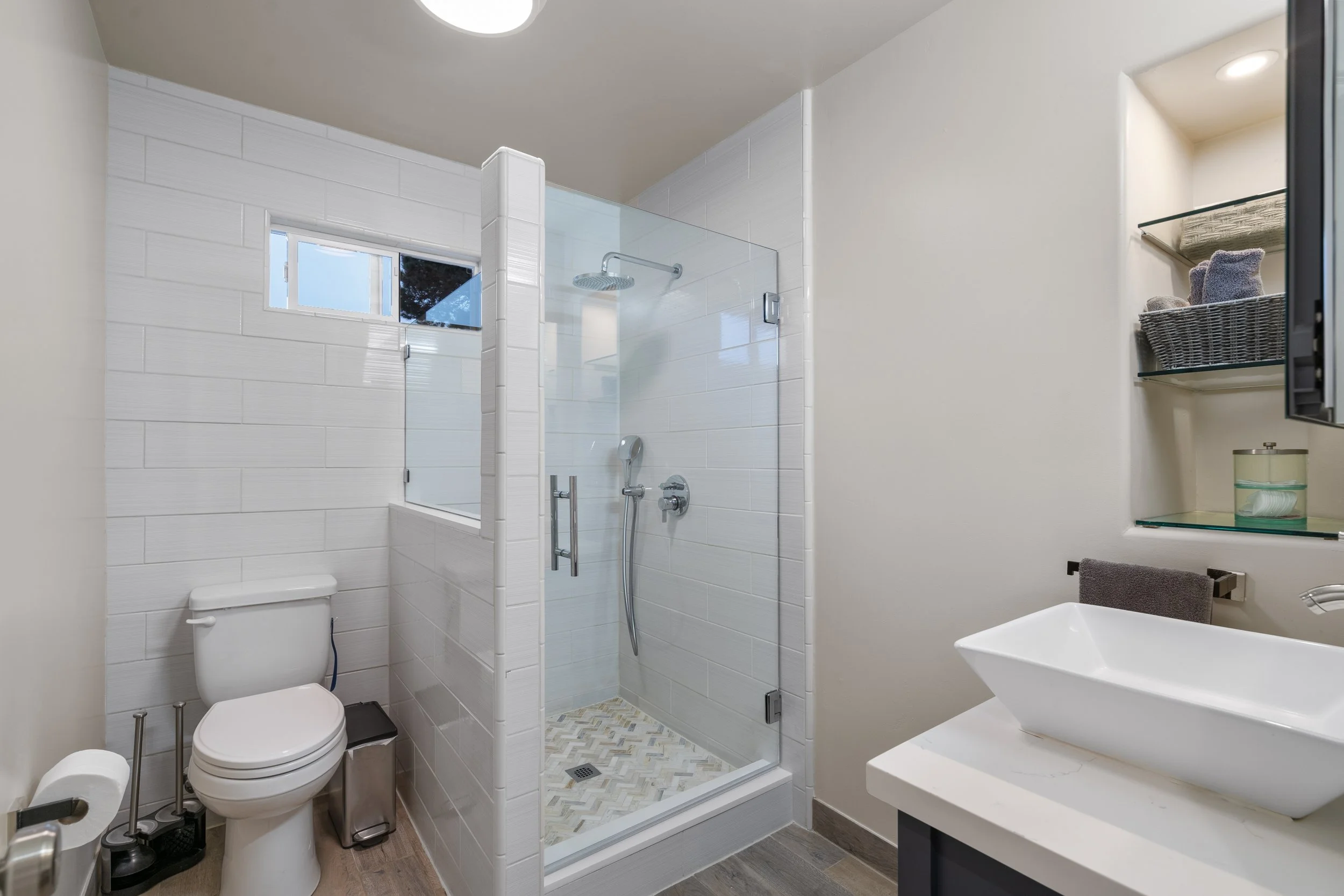 A modern bathroom with a glass-enclosed shower, white walls, a toilet, a small window, and a white vessel sink on a countertop.
