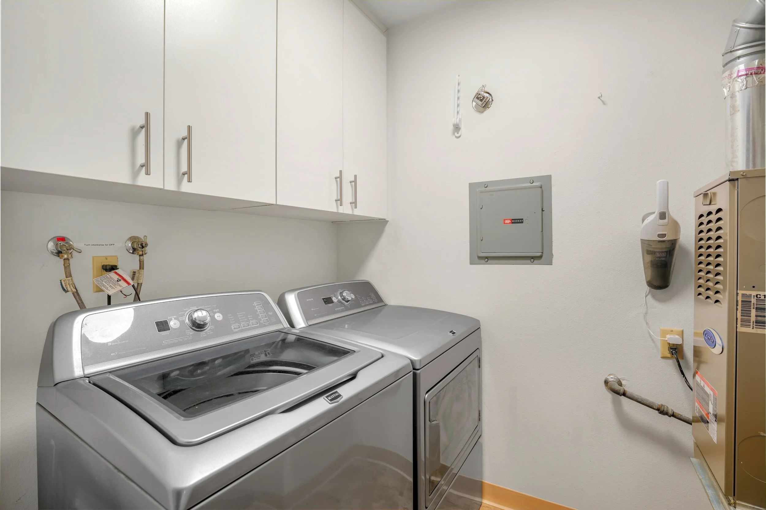 A laundry room with a top-loading washing machine and a front-loading dryer, white cabinets above, and utility connections on the wall.