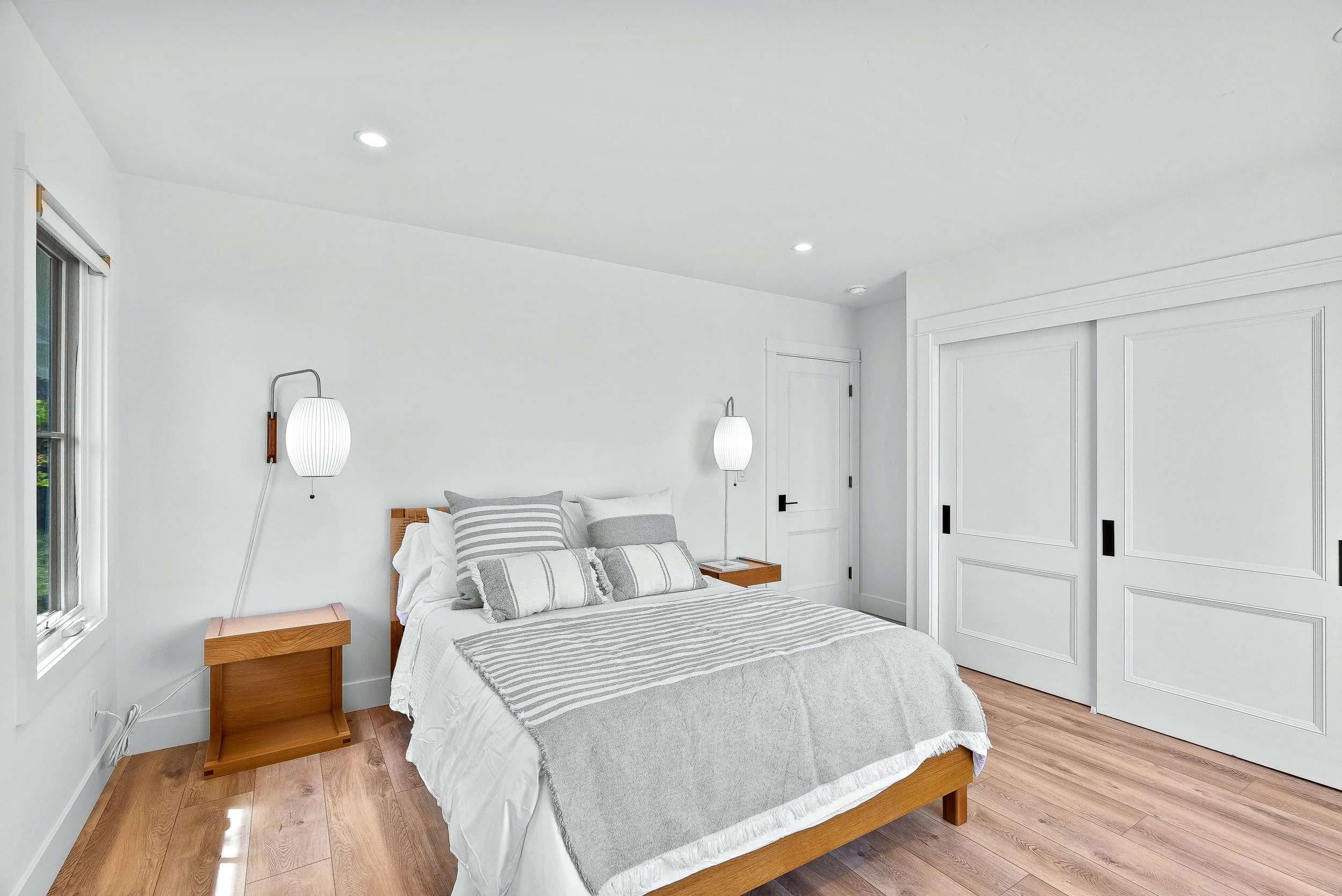 A bedroom with a wooden bed frame, white bedding, and gray striped pillows, featuring two wall-mounted lamps and a closet with sliding door in Capitola, California.
