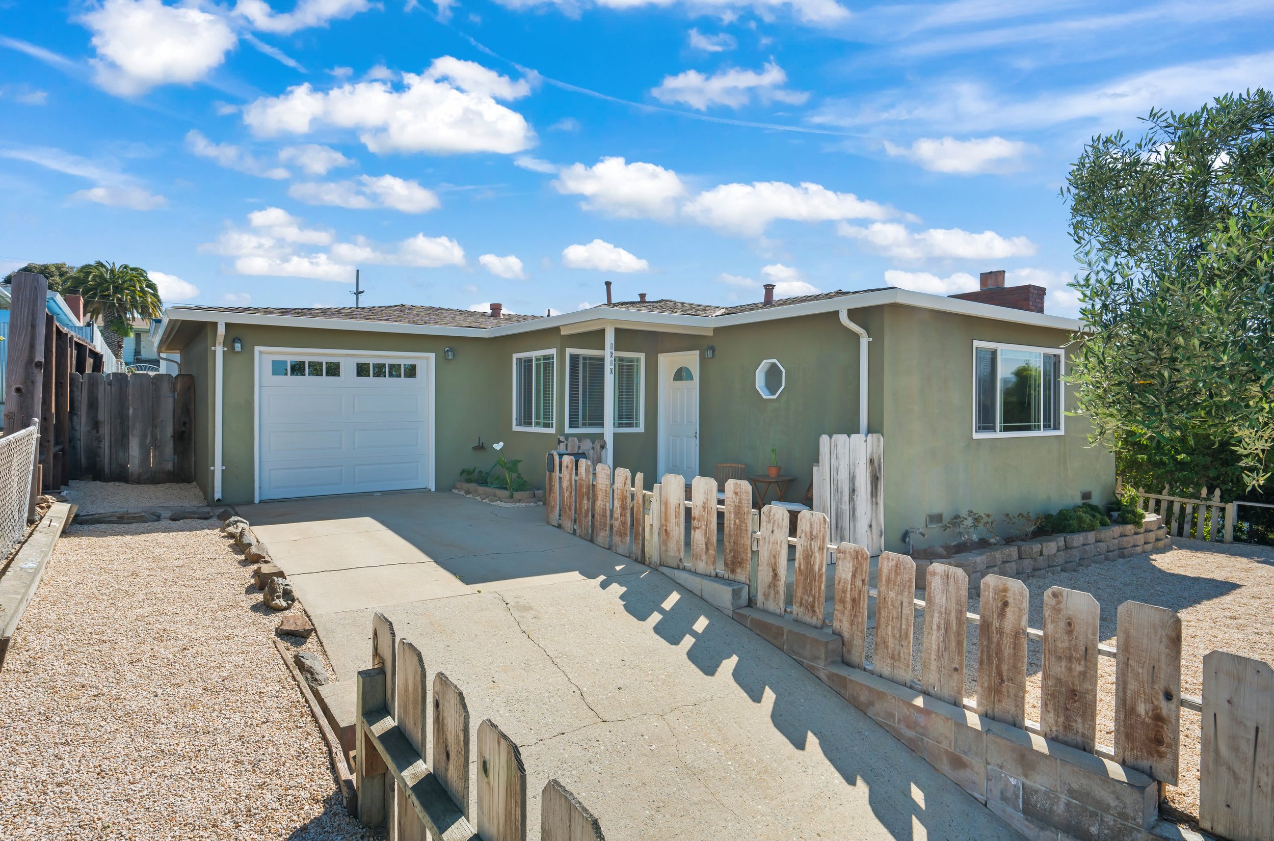 Front view of a single-story house with a driveway, green exterior, white garage door, and small garden bed with plants and a star-shaped decoration. Wooden fence surrounds part of the yard, and there are large windows and a small door. The sky is blue with scattered clouds.