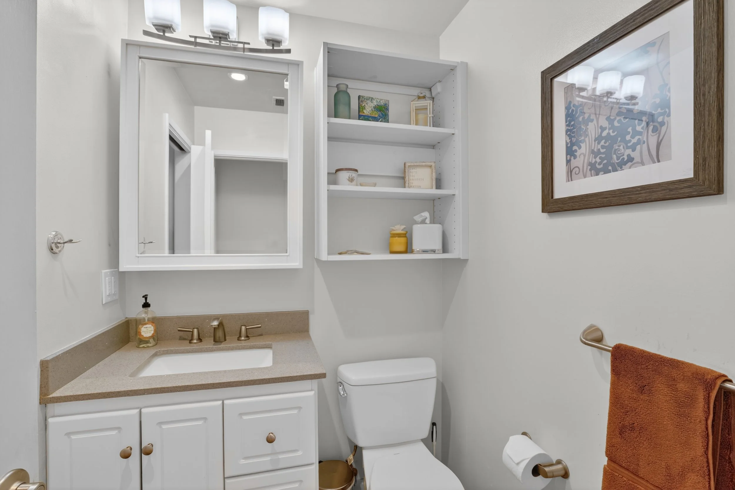 A small bathroom with a white vanity, beige countertop, and a medicine cabinet mirror above. There is a white toilet and a wall-mounted shelf with decorative items. A framed artwork is on the wall, and towels are on a rack.
