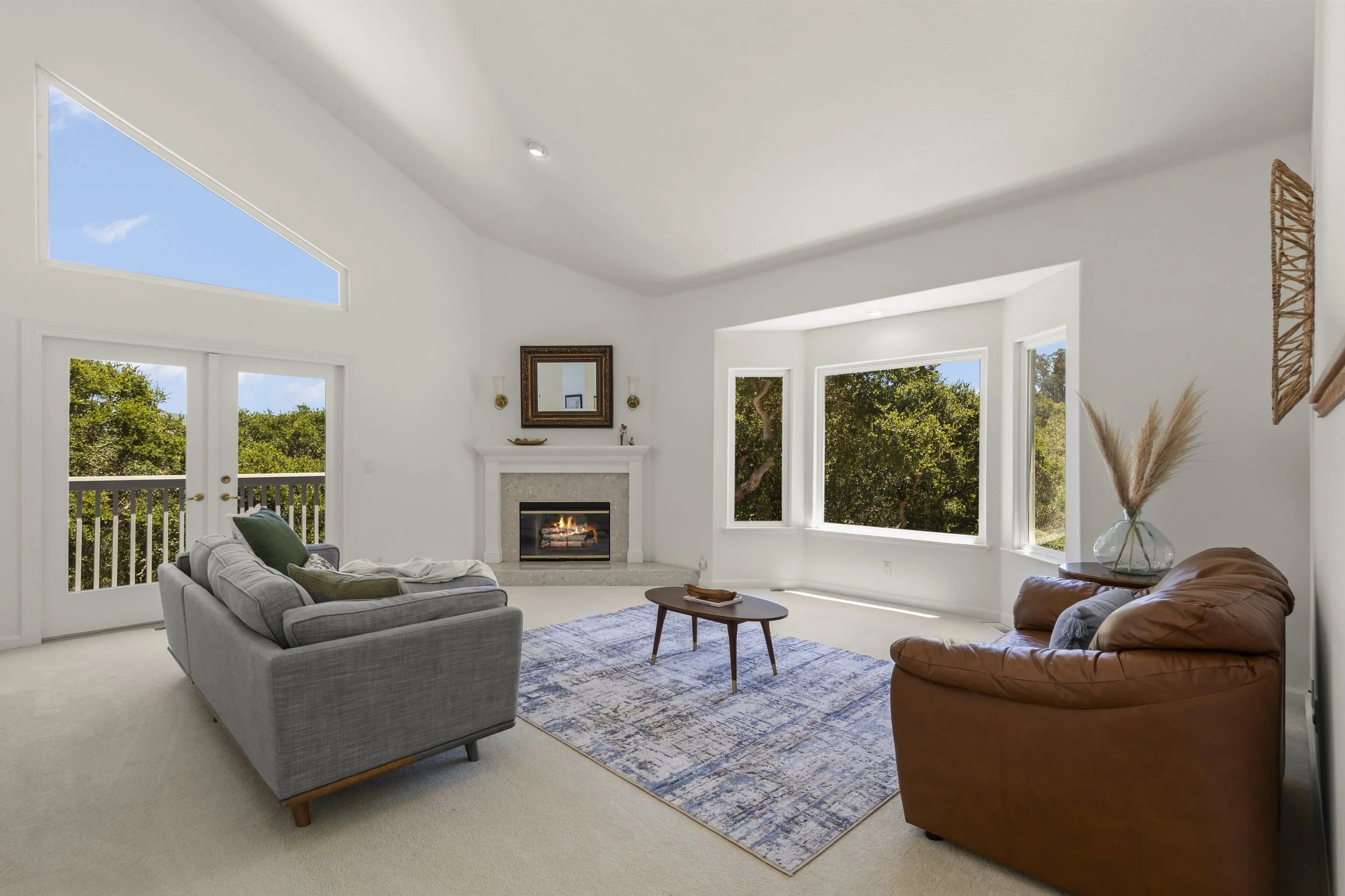 Bright living room with large windows, a fireplace, a gray sofa, a brown leather armchair, and a small wooden coffee table on a patterned rug.