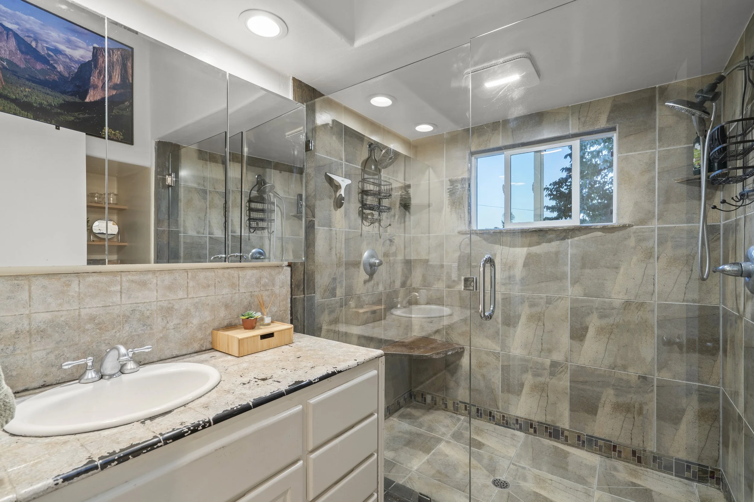 Bathroom with a glass-enclosed walk-in shower, beige tile walls, a small window, a white vanity with a granite countertop, a small plant in a container, and a mirror.