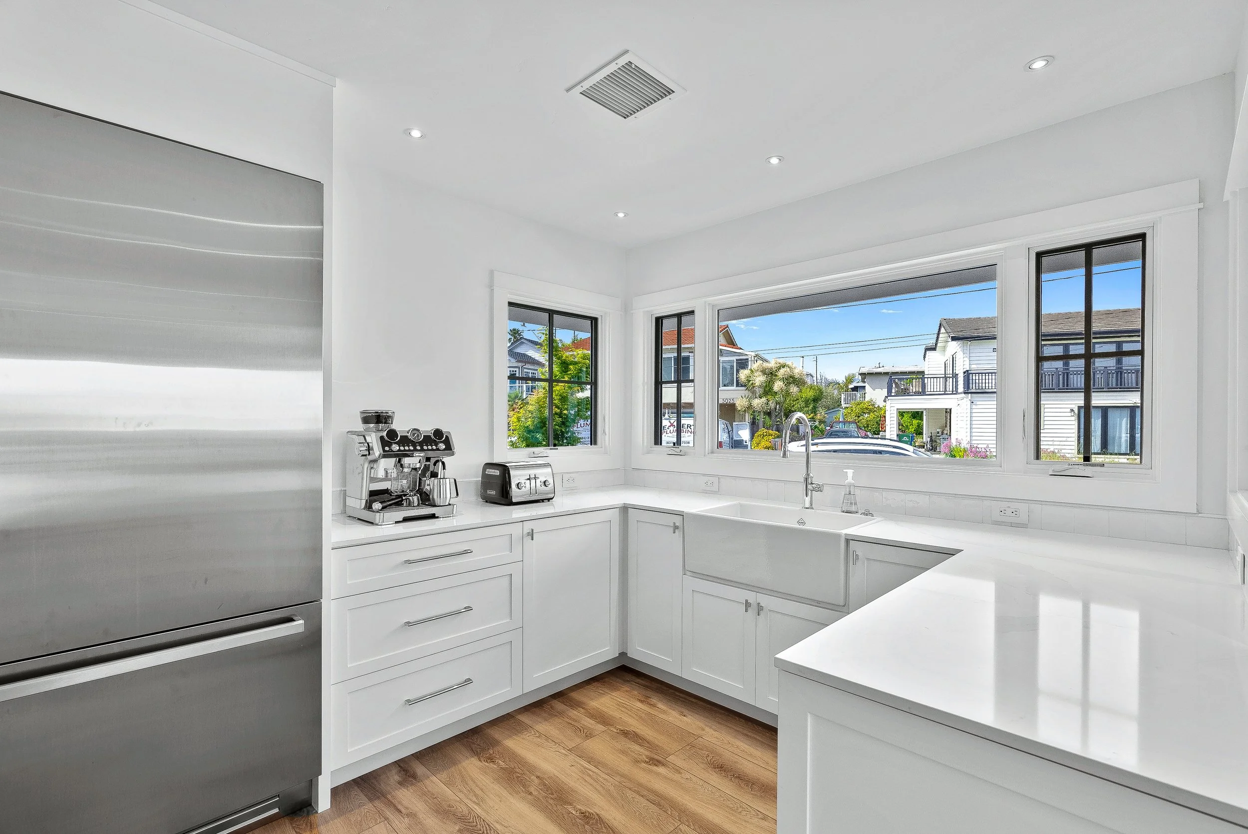 Modern kitchen with white cabinets, stainless steel refrigerator, coffee machine, toaster, front-facing sink, and large windows overlooking neighboring houses and blue sky. in Capitola, California.